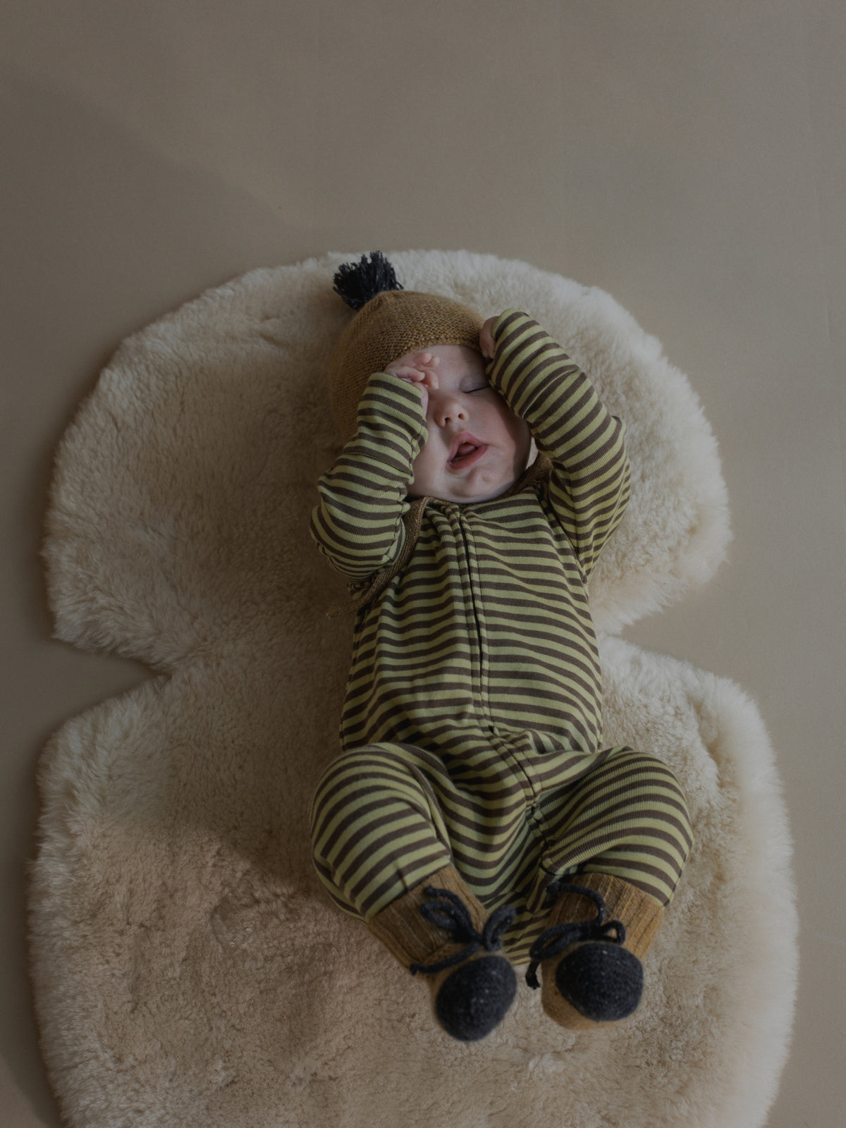 A baby lying on a cream sheepskin rug, wearing a green and olive striped footed one-piece with a tan hat and dark booties.