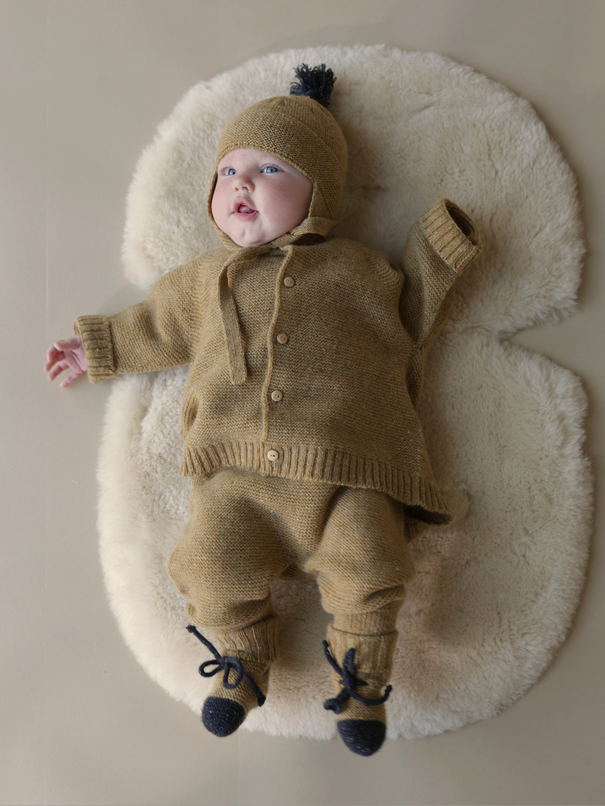 A baby wearing a tan knit three-piece outfit with a matching bonnet and dark blue booties, lying on a white sheepskin rug against a neutral beige background.