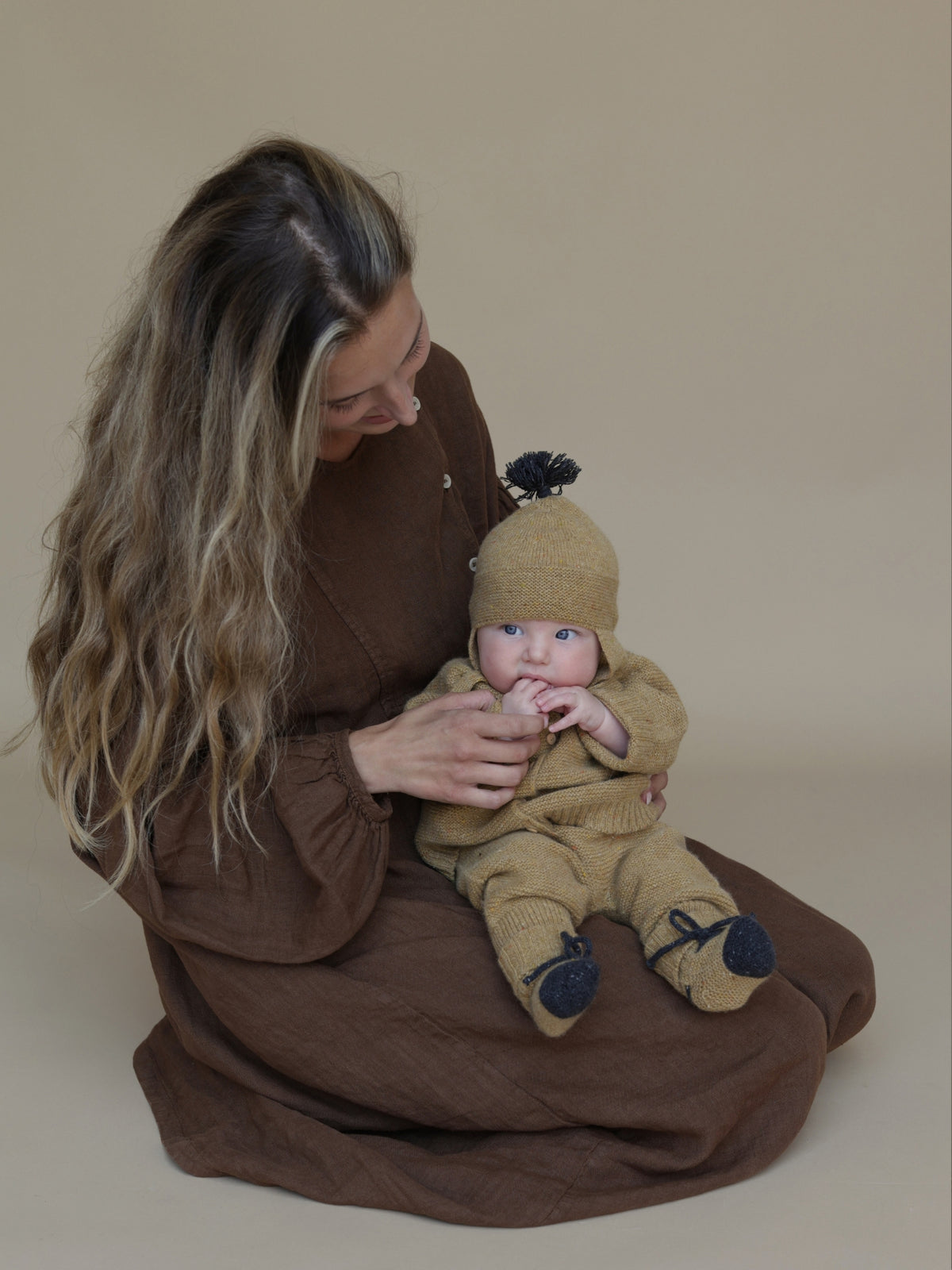 A woman in a brown dress sits on a beige studio floor holding a baby wearing a matching tan knit bonnet and footie suit with dark pom-poms and booties. Model is 62cm and wears 0-3m