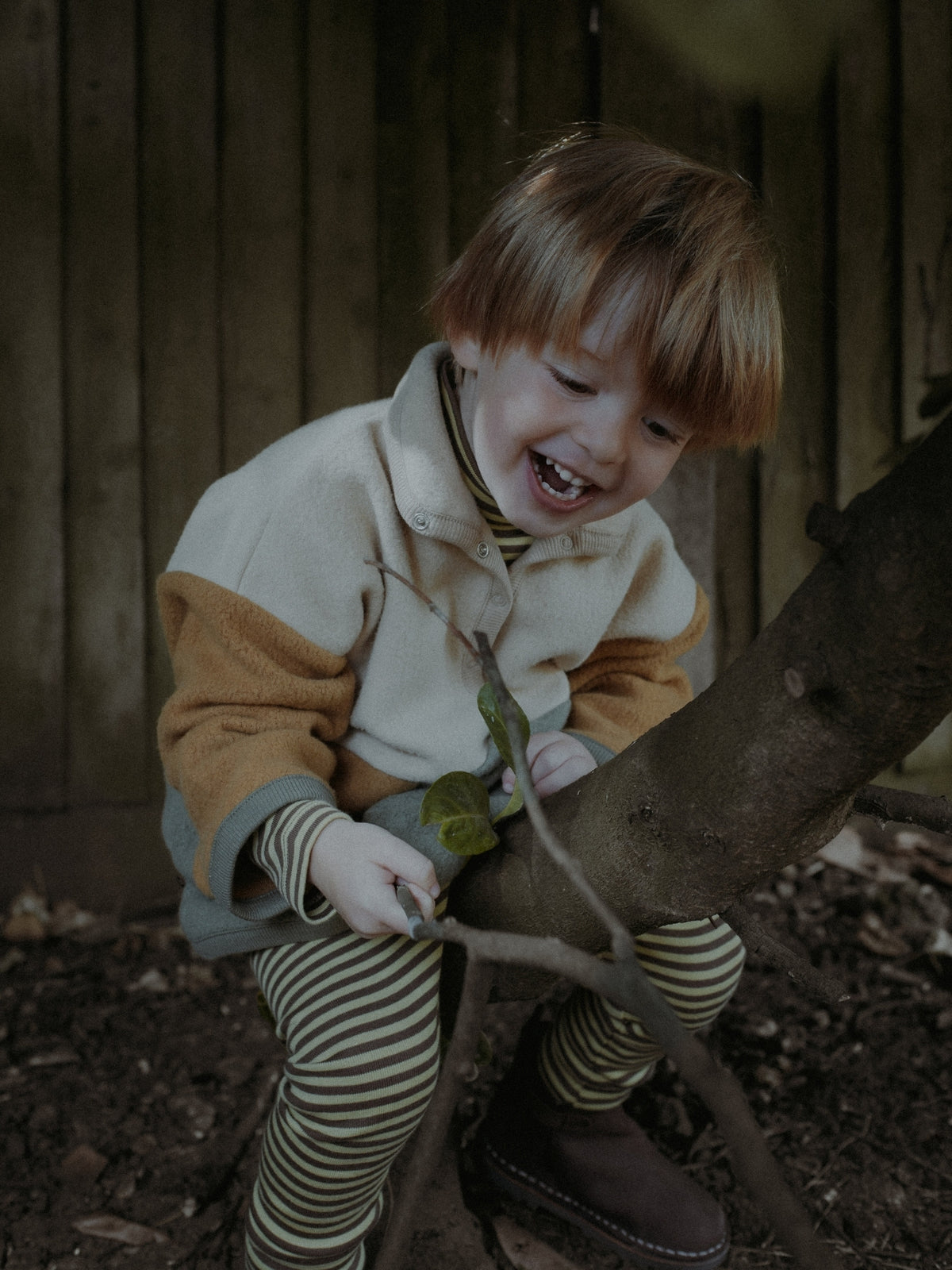 A young child with light brown hair laughing while sitting on a tree trunk outdoors, wearing a cream and tan color-blocked fleece jacket, striped green and beige leggings, and brown boots against a dark wooden fence background.