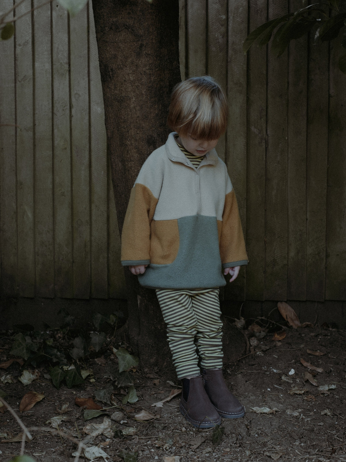 A toddler stands outdoors against a wooden fence and tree trunk, wearing a color-block fleece pullover with cream, mustard, and muted blue-green panels, striped leggings, and dark rain boots.