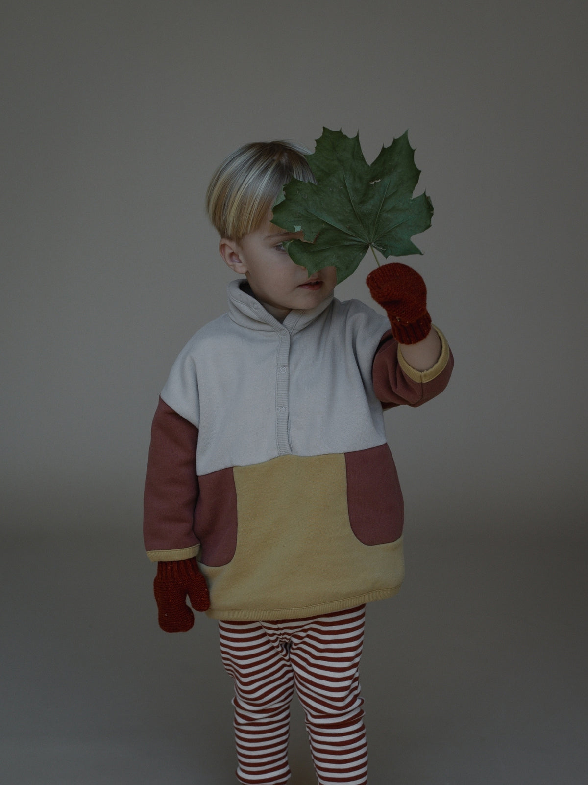 A child wearing a color-block fleece jacket in white, brown, and mustard yellow, paired with red and white striped pants and red mittens, holding a green leaf in front of their face against a plain gray studio background.