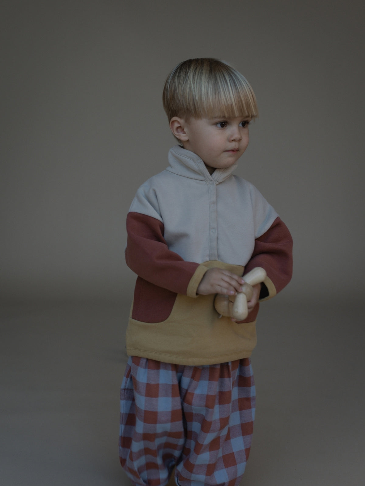 A toddler standing in a studio wearing a color-block fleece jacket with a light gray upper section, dark red sleeves, and a tan front pocket, paired with red and blue plaid pants.