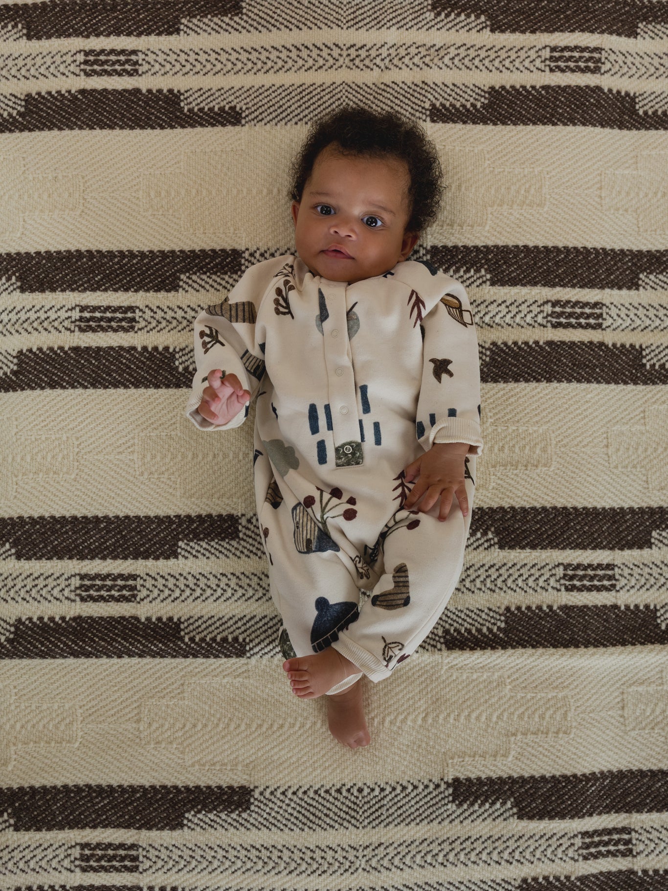 baby in cream undyed cotton onesie with cosy nature-themed hand-drawn prints, lying on carpet