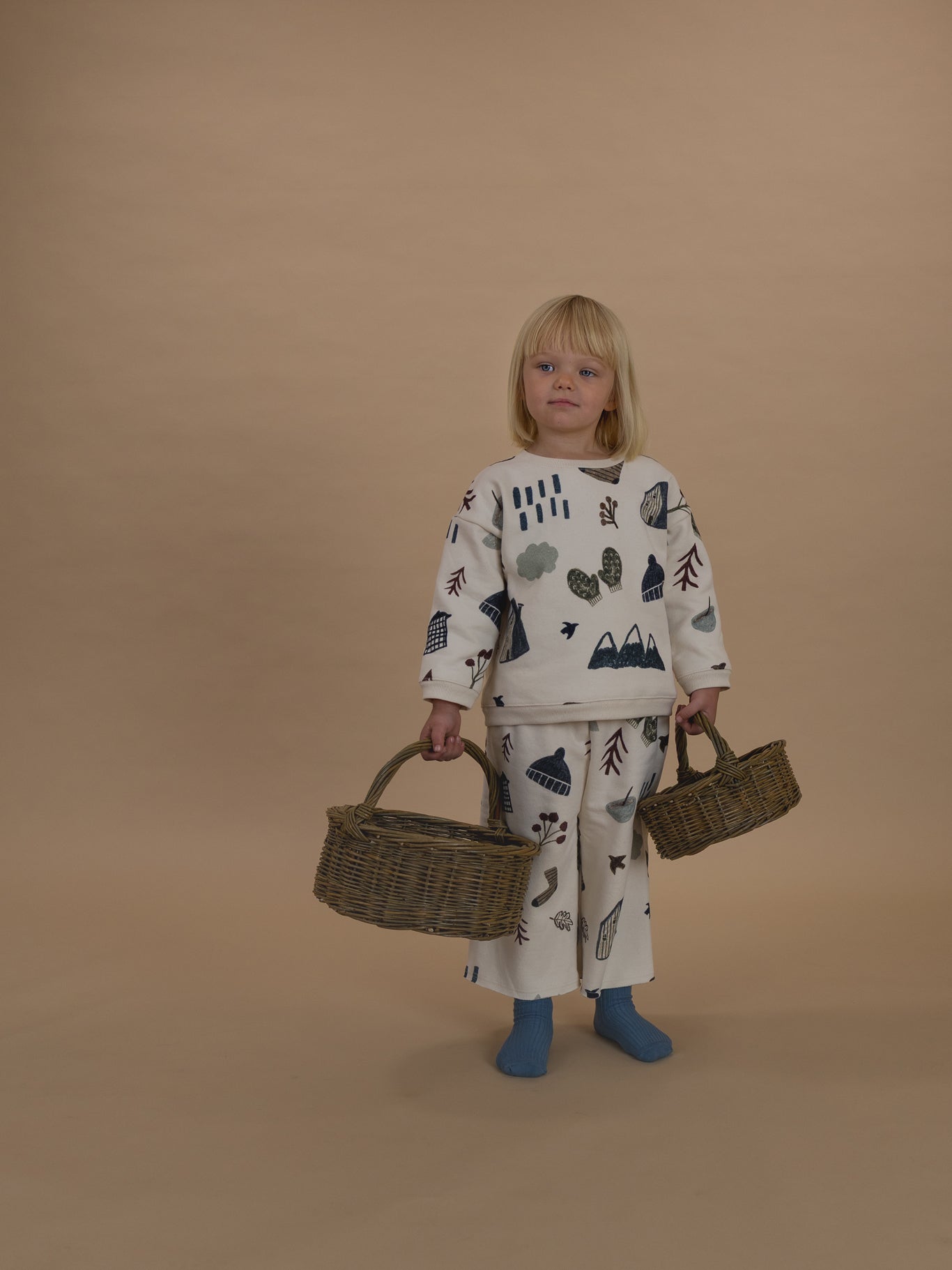 toddler in matching cream cotton sweatshirt and pants with cosy nature-themed graphics, holding baskets
