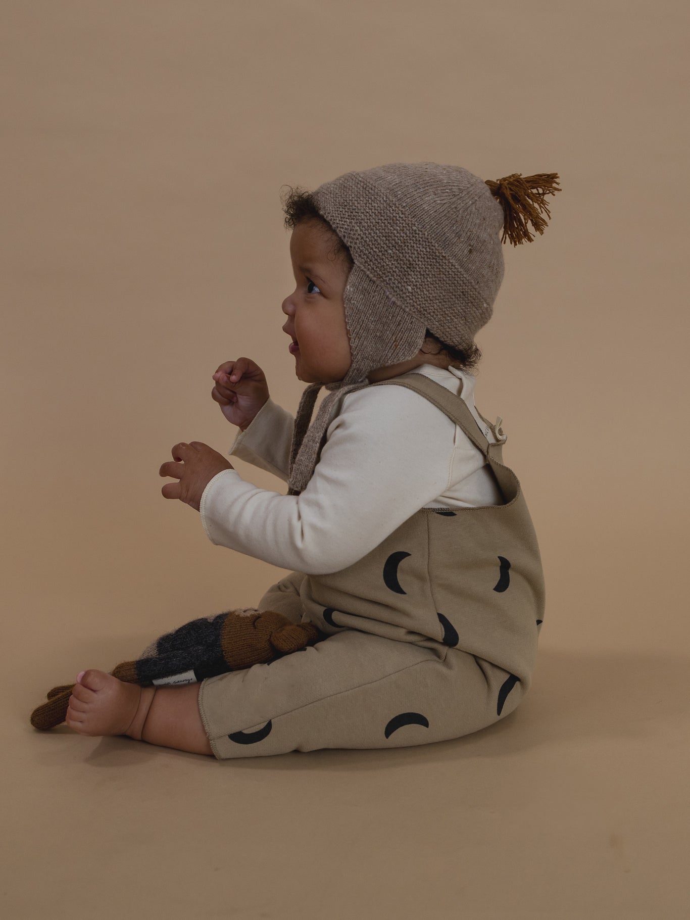 side of baby in dusky beige salopette with black moon print, white sweatshirt & oatmeal wool tassel hat