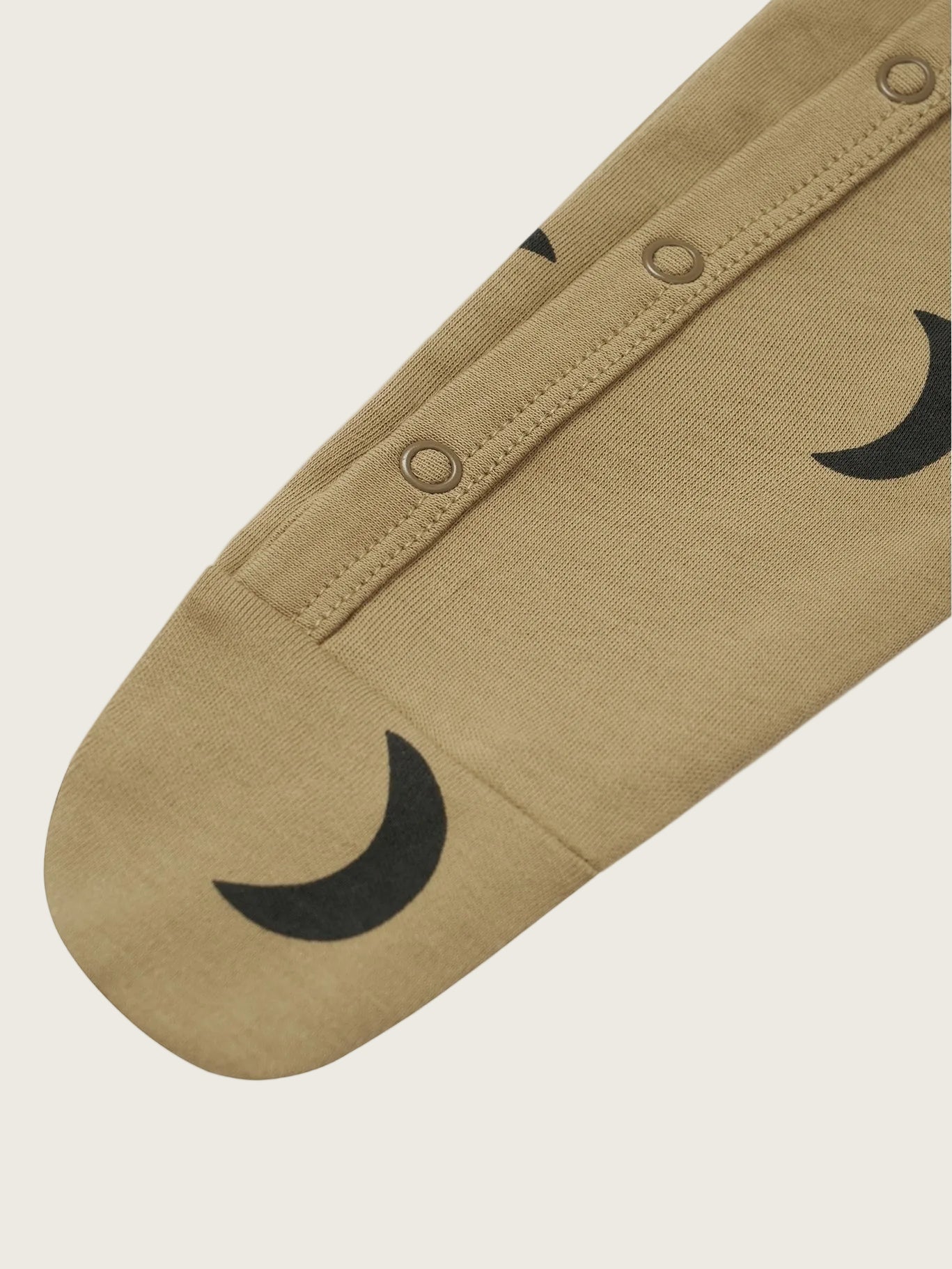 A close-up of a beige fabric garment, likely a sleeve or leg warmer, featuring a black crescent moon pattern and three metal snap buttons along a seam, set against a plain white background.