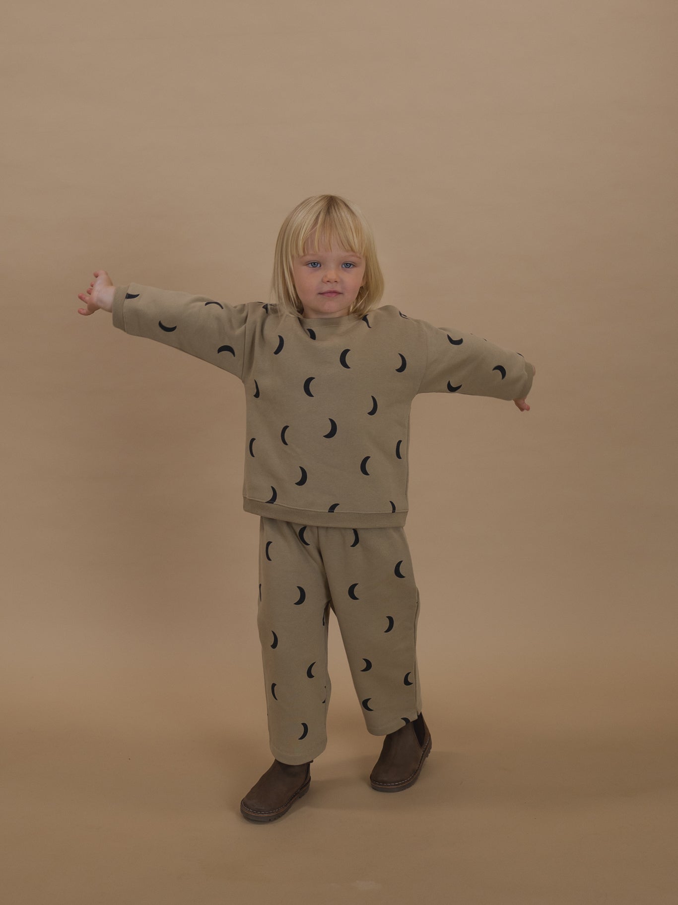 toddler in dusky beige sweatshirt with black moon print & matching pants, standing with arms out