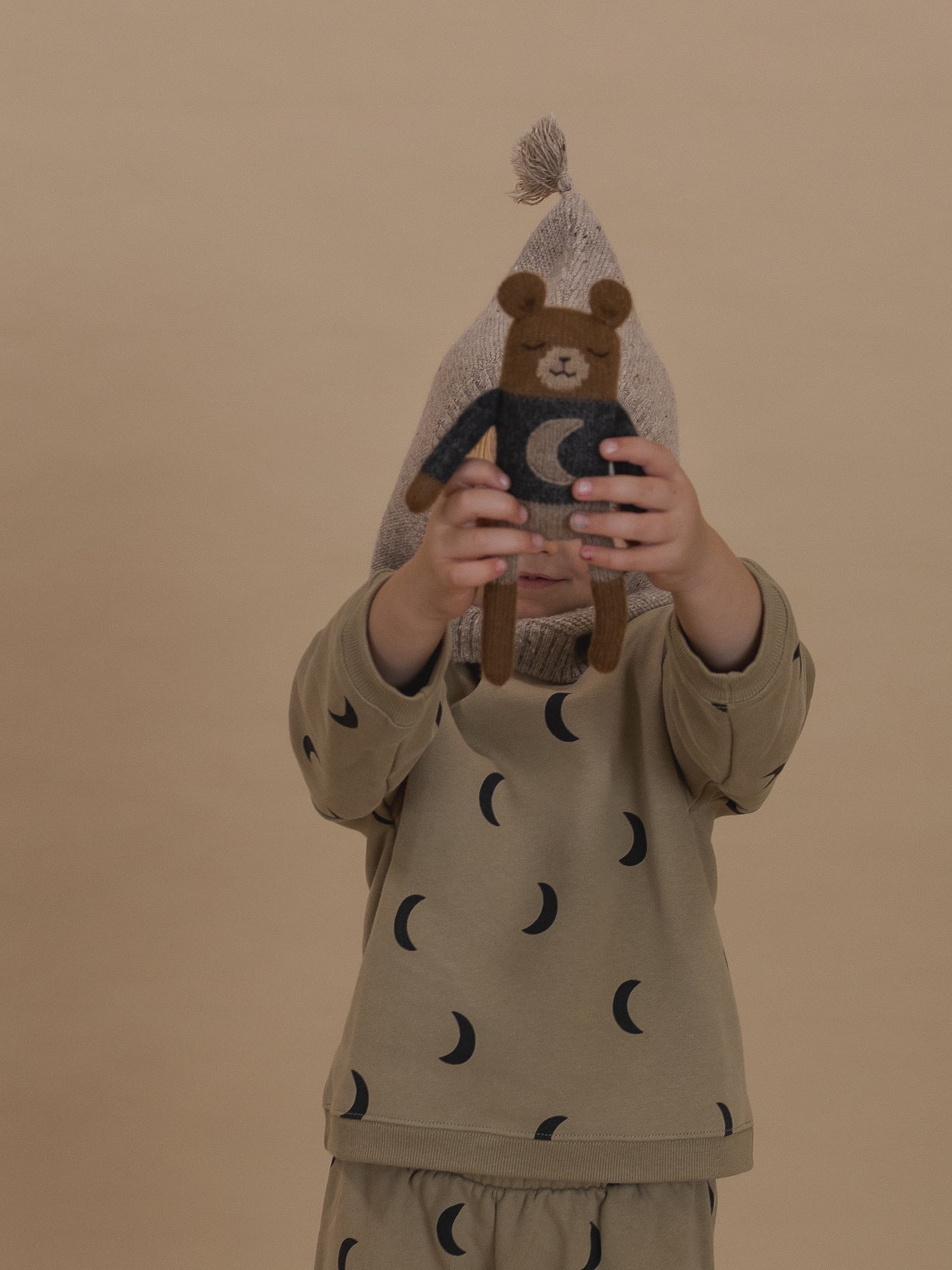 toddler in beige sweatshirt with black moon print & matching pants, holding a wool teddy plushie