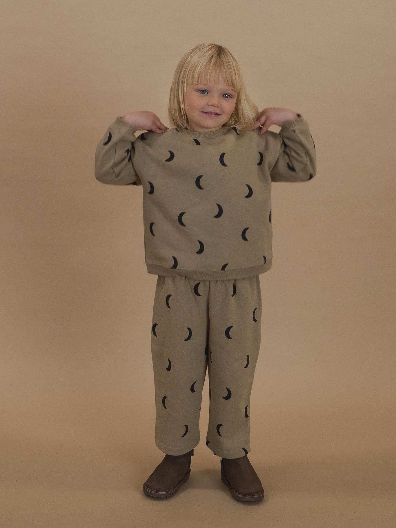 toddler in dusky beige trousers with black moon print and matching sweatshirt, hands on shoulders