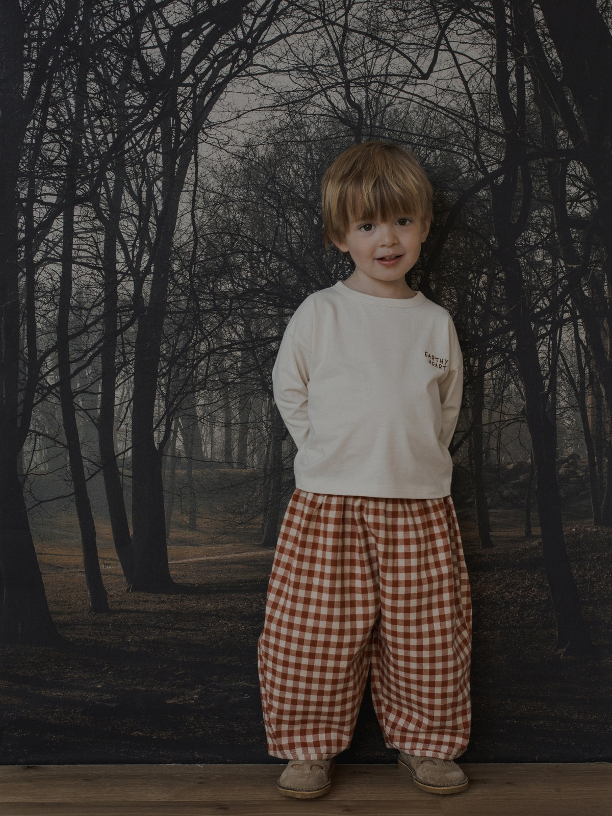 A young child stands in front of a dark forest backdrop, wearing a cream long-sleeve shirt and loose red-and-white gingham pants.
