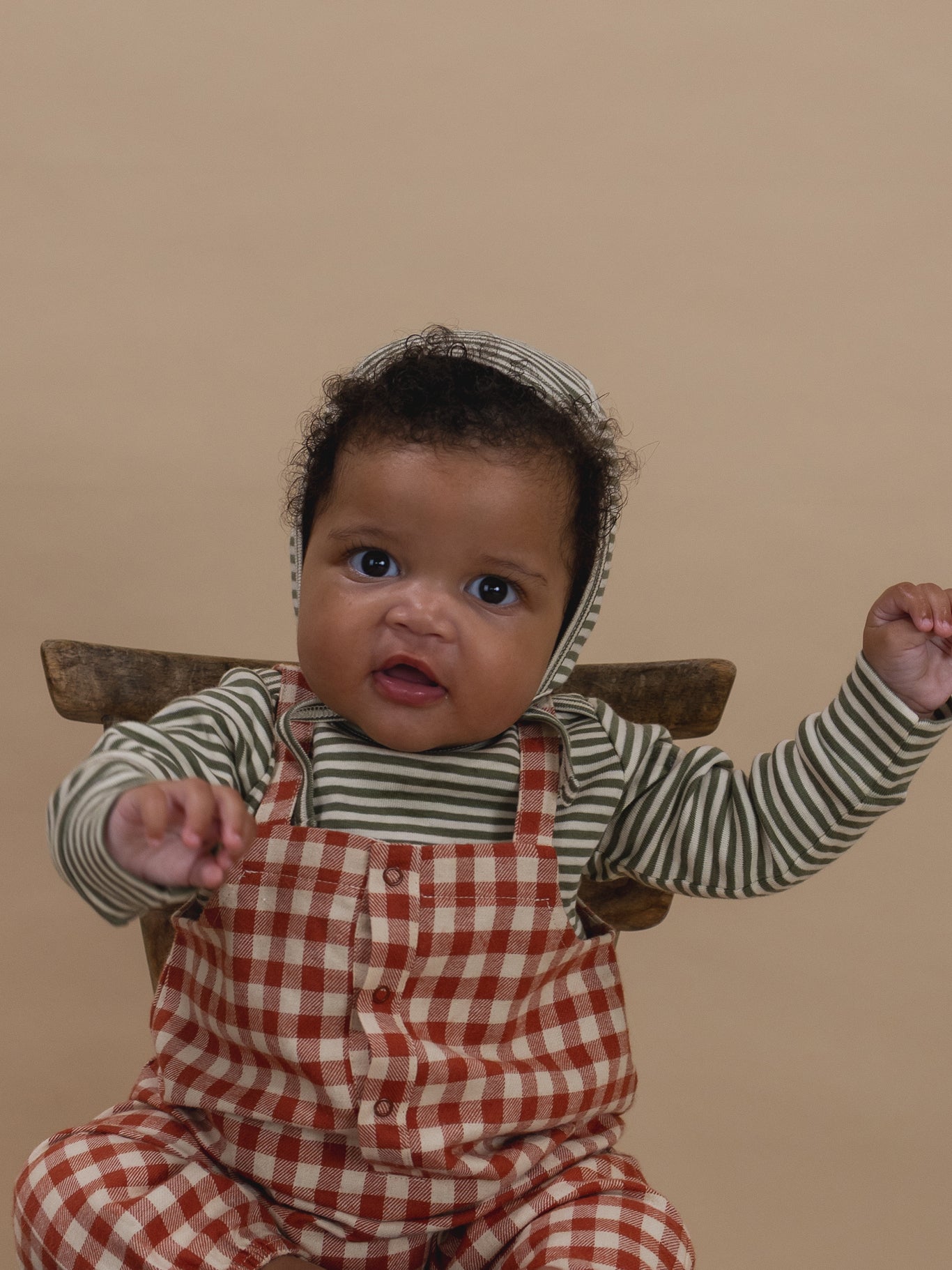 Baby wearing a striped shirt with checkered overalls, and fern green striped bonnet, sitting on chair