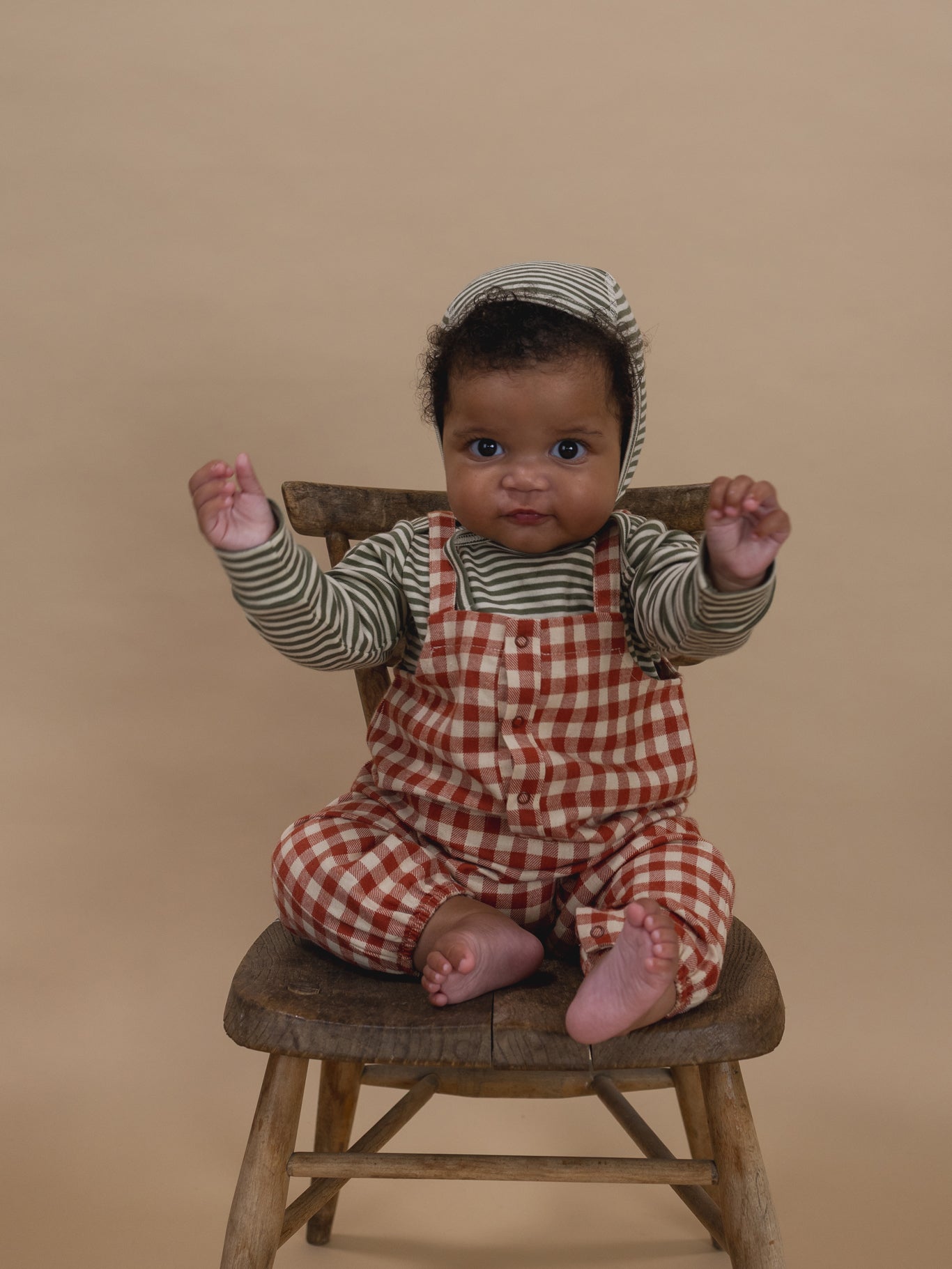 Baby sitting on a wooden stool wearing a checkered outfit and fern green and white stripes bonnet