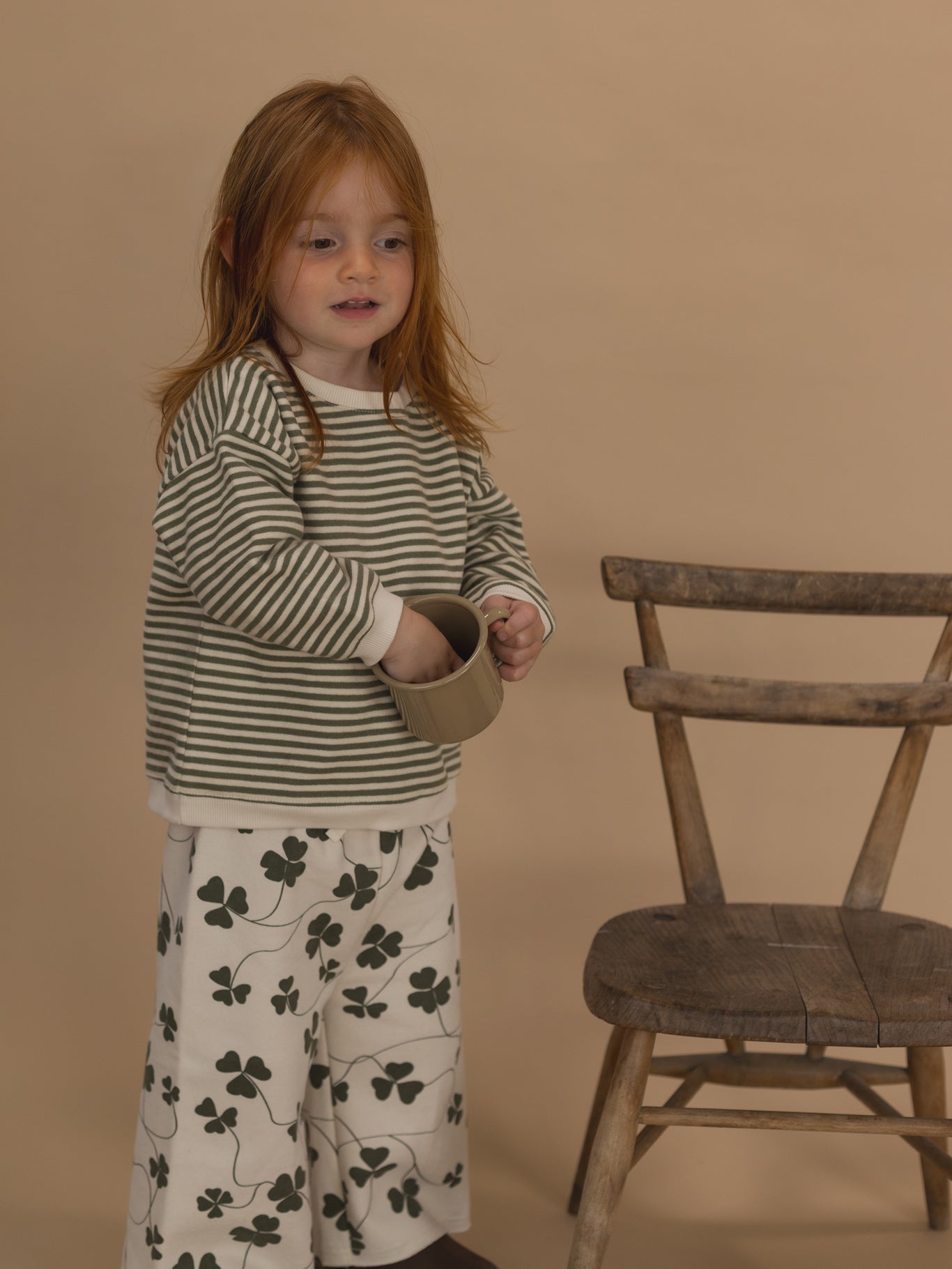 toddler in green and white striped sweatshirt, and wide leg pants with clover print, holding a cup