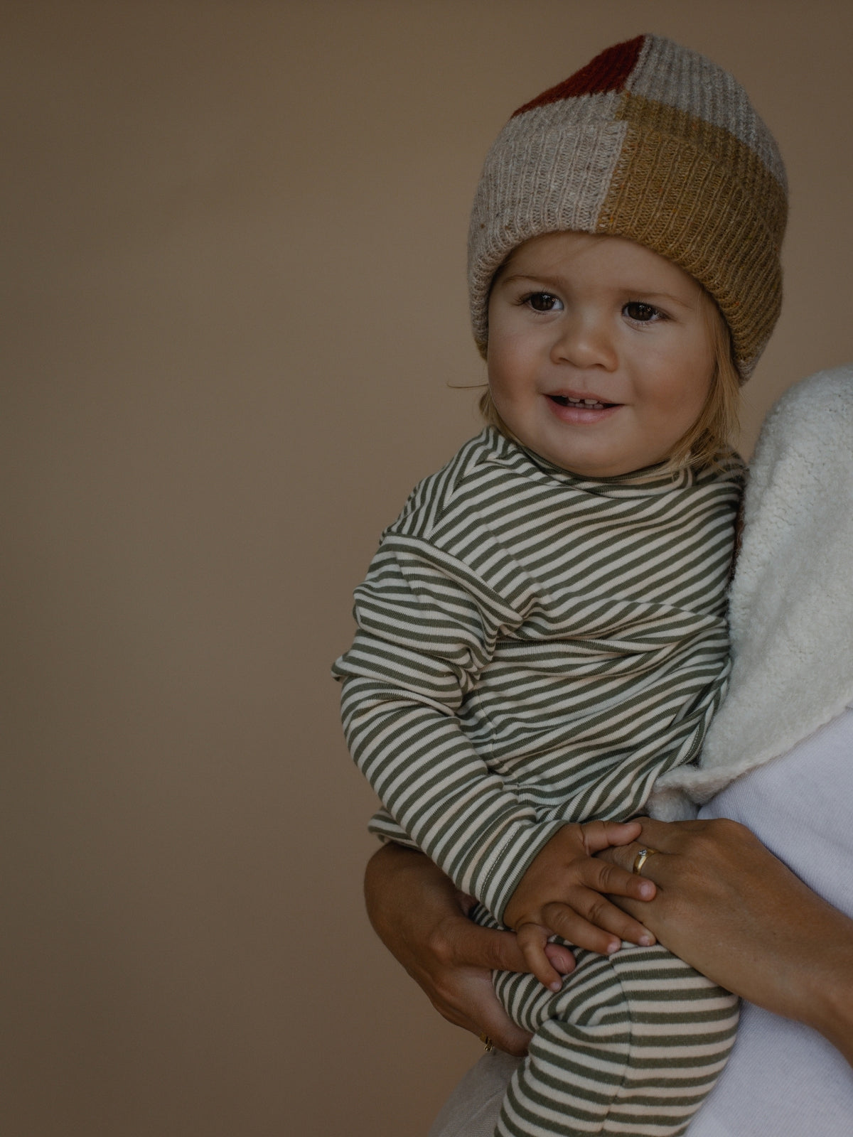 A baby wearing a beige and brown striped long-sleeve bodysuit and a chunky knit patchwork beanie with beige, brown, cream, and red sections, held by an adult against a solid beige background.