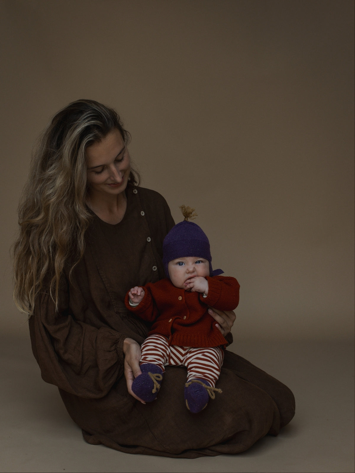 A woman in a brown dress sits on the floor holding a baby wearing a red knit cardigan, striped pants, and a purple hat against a plain brown studio background.