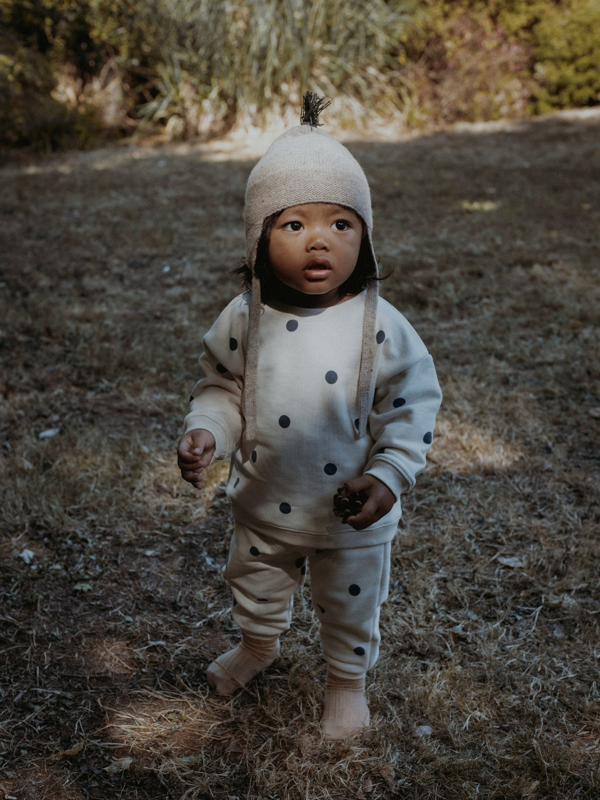 A toddler standing outdoors on dry grass wearing a cream knit one-piece outfit with black polka dots, a matching knit bonnet with a small topknot, and light socks.