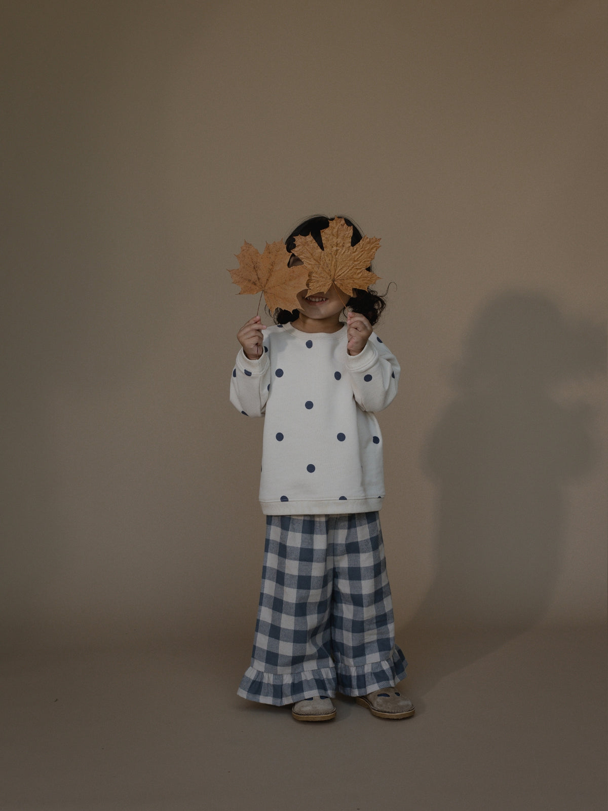A child stands against a plain warm beige studio background holding two large brown paper maple leaves in front of their face. The child is wearing a white long-sleeve top with dark blue polka dots and blue-and-white gingham wide-leg pants.