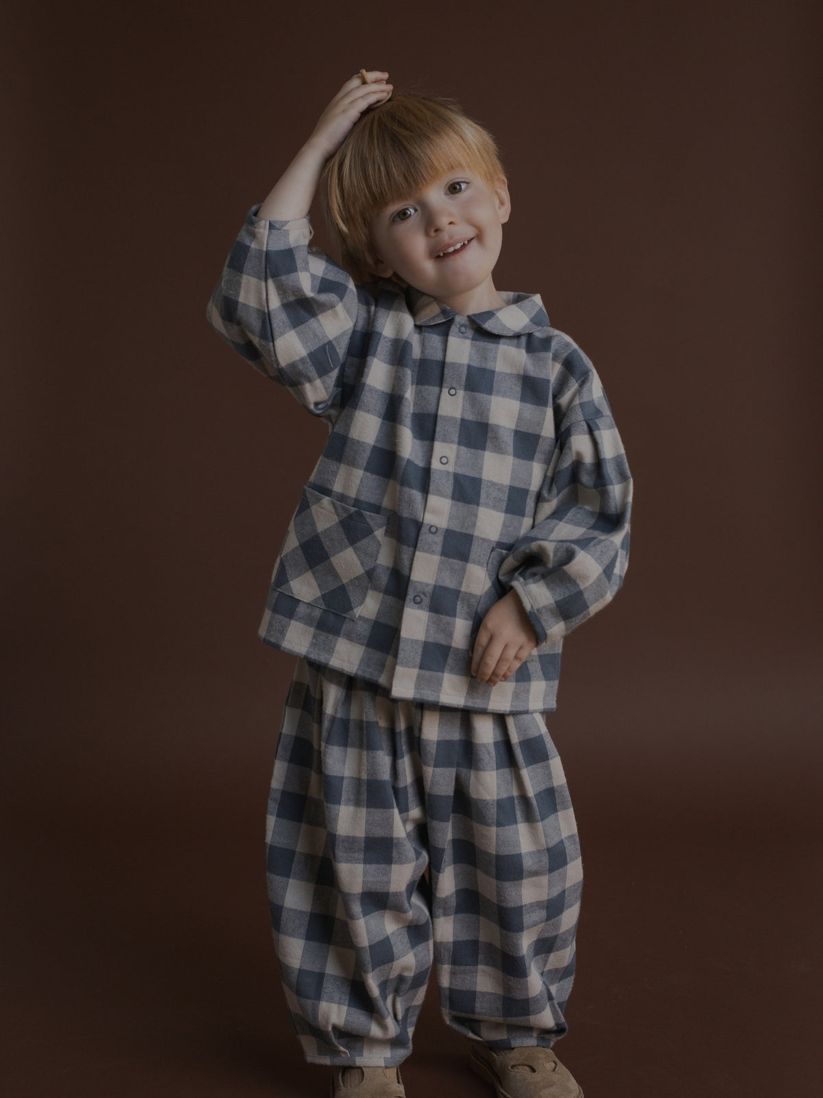 A young child wearing a matching two-piece coord with a blue and beige gingham check pattern, standing against a solid dark brown studio background.