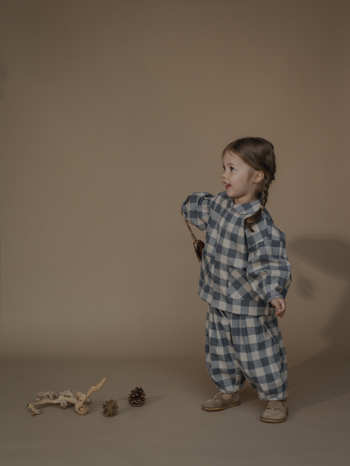 A young child wearing a matching blue and white checkered pyjama set stands against a plain beige studio background. The set includes a long-sleeve top and loose pants, styled with beige shoes and a small brown shoulder bag.