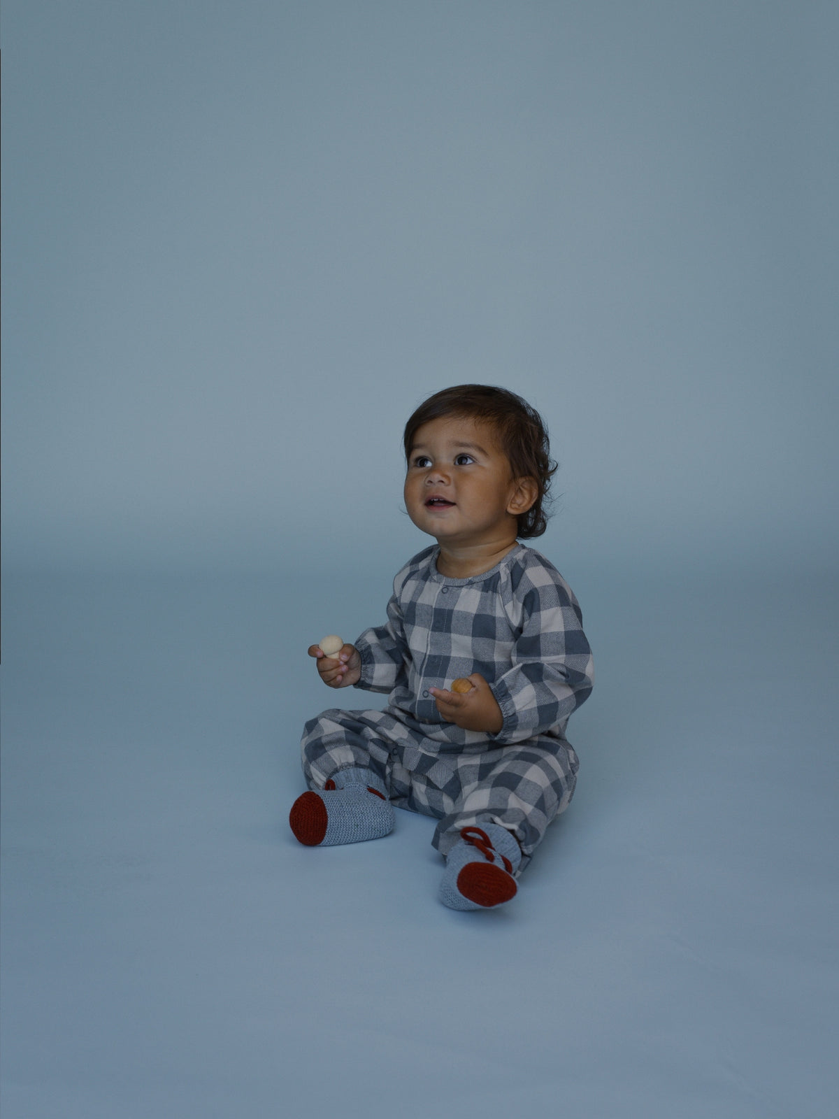 A baby sitting on a light blue studio background wearing a gray and white gingham check one-piece set with red socks.