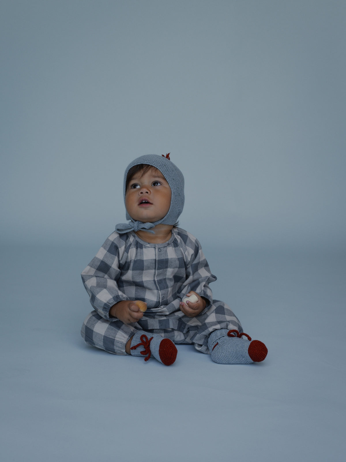 A baby sitting on a light blue-gray studio background, wearing a matching plaid hooded romper and red booties, holding small wooden and white objects.