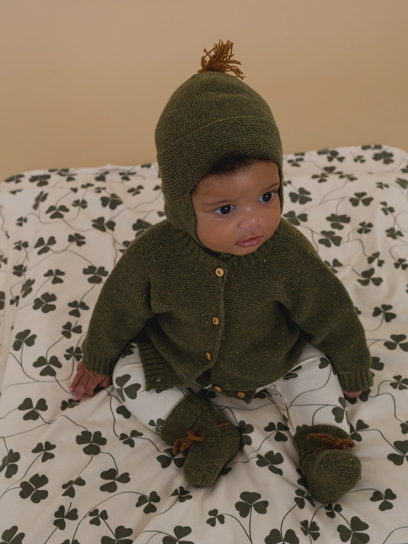 baby in forest green wool hat with brown tassel, matching cardigan and booties, clover sleepsuit