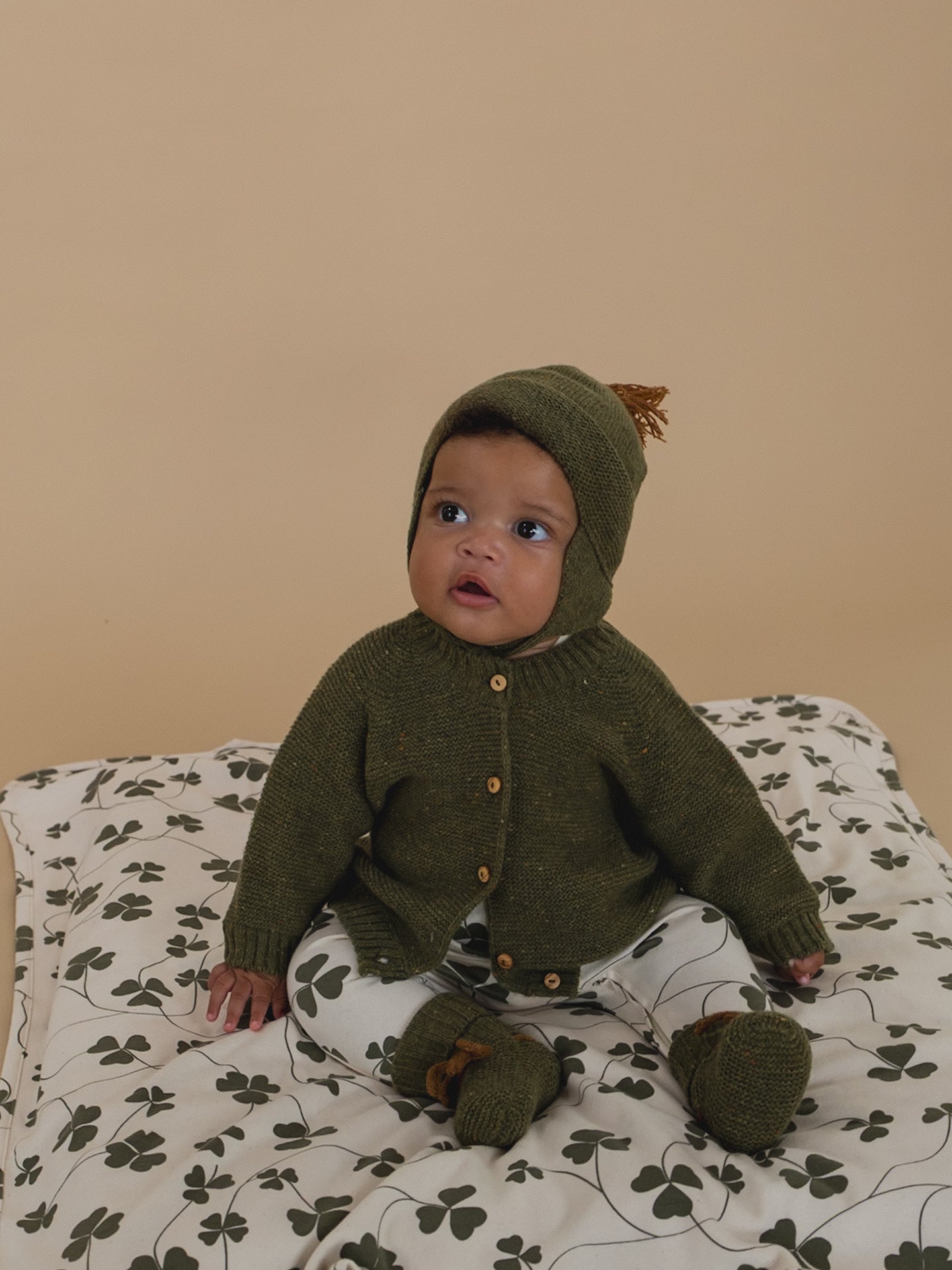 Baby wearing a green knitted outfit with a hood on a clover pattern blanket.