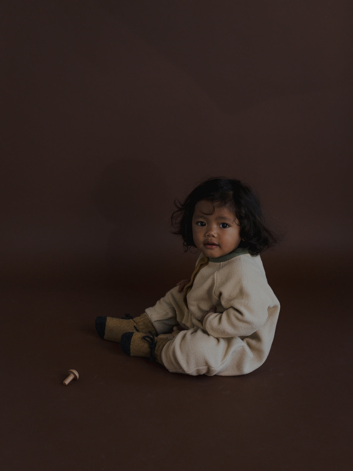 A toddler with dark curly hair sits on a dark brown studio floor wearing a cream-colored knit romper and tan socks.