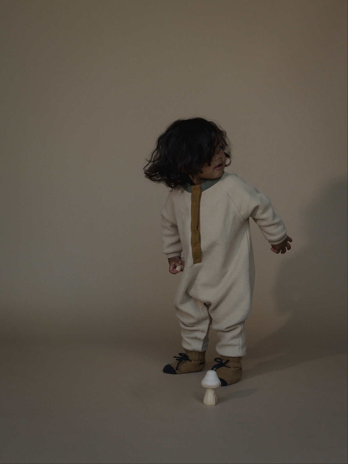 A toddler stands in profile wearing a cream-colored one-piece jumpsuit with a vertical mustard yellow zipper detail and dark brown boots, set against a plain beige studio backdrop.