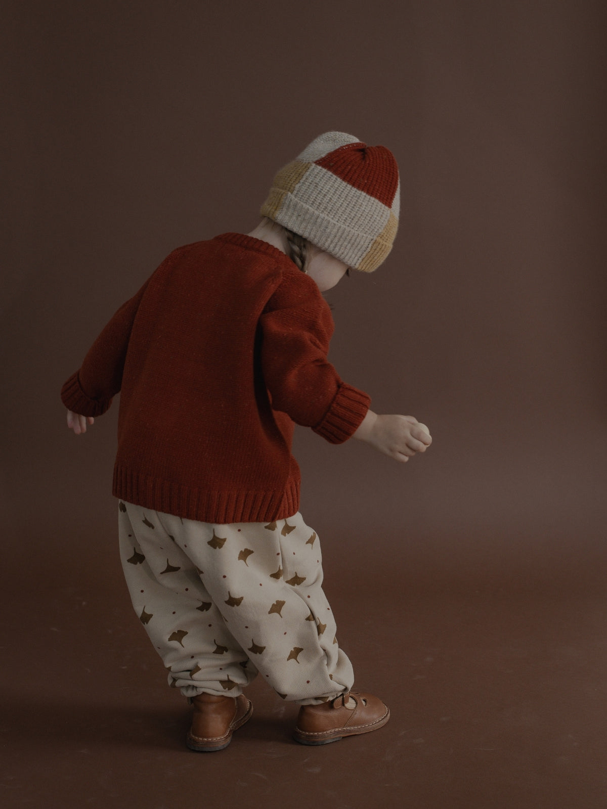 A toddler standing with their back to the camera, wearing a rust red knit sweater, cream pants with a small brown pattern, and a red, cream, and tan knit beanie against a solid dark brown studio background.