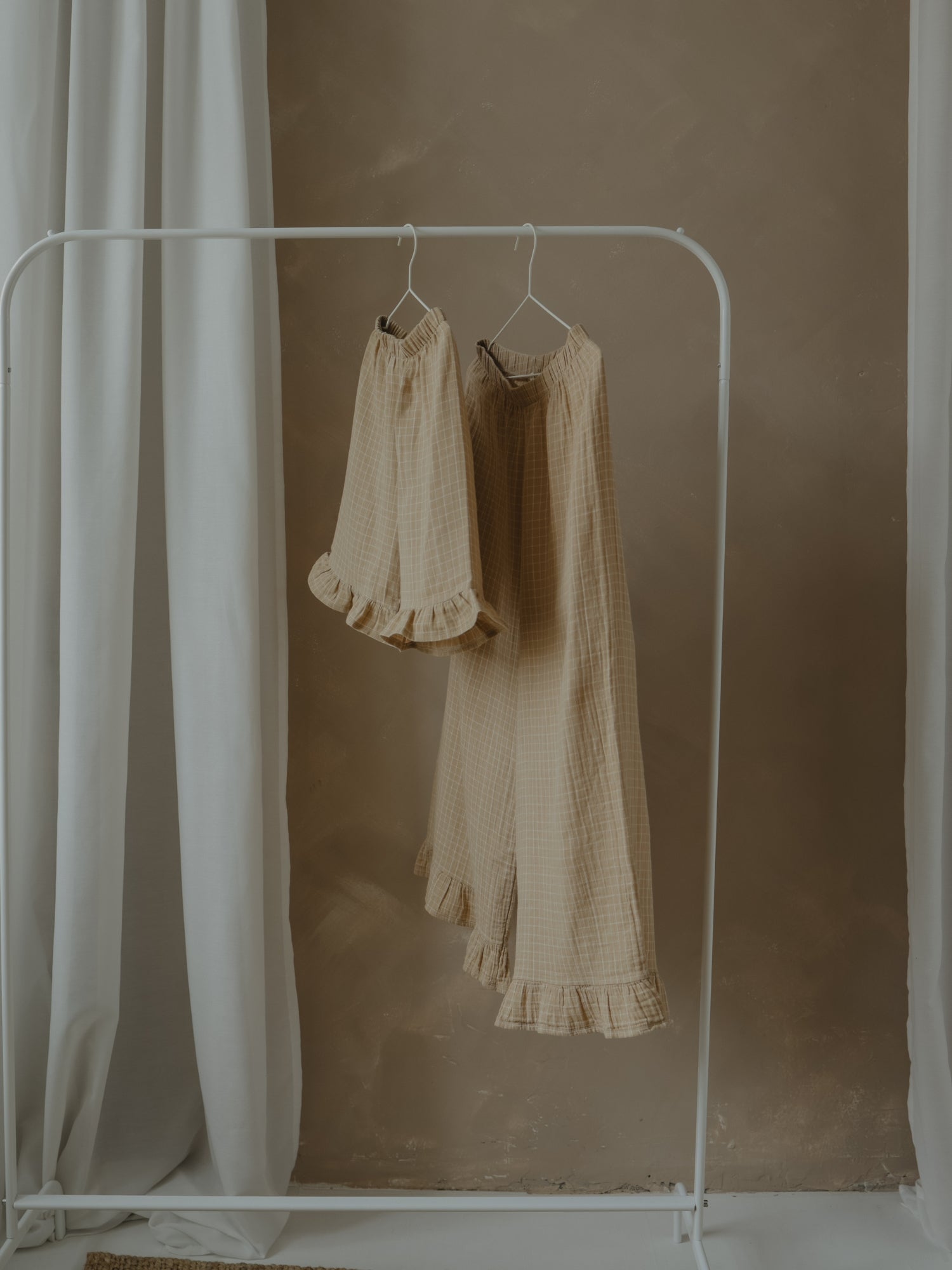 Two beige bottoms hanging on a white rack against a plain wall.