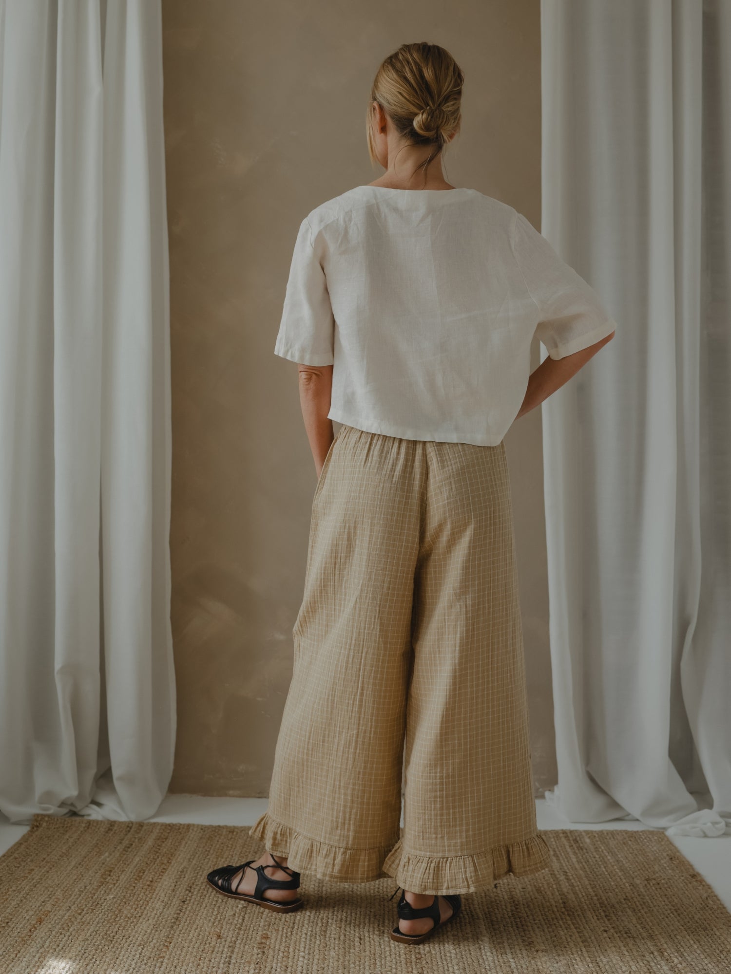 Person wearing a white top and beige checkered pants standing in front of white curtains.