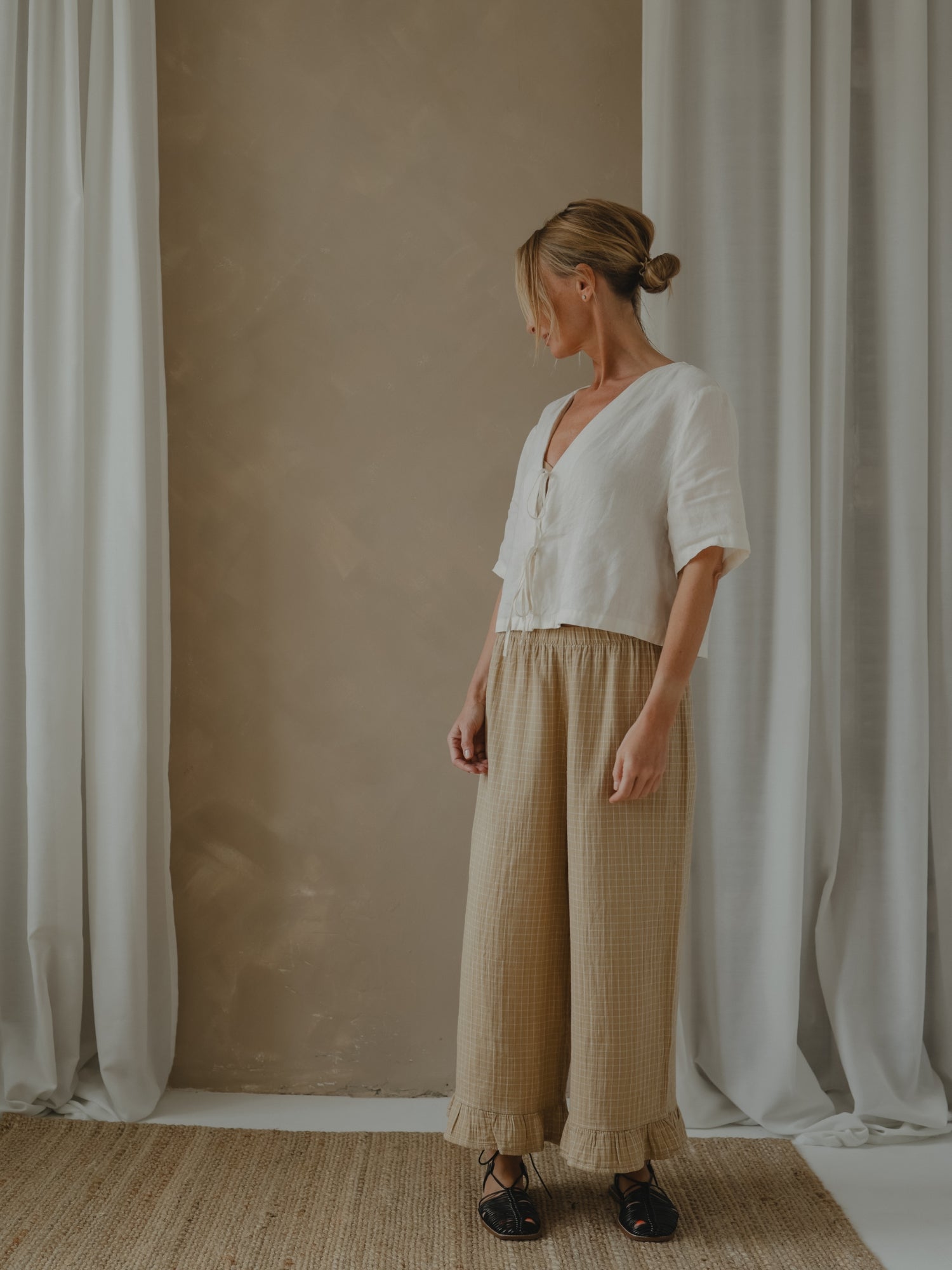 Woman wearing a white blouse and beige pants standing in front of a beige wall with white curtains.