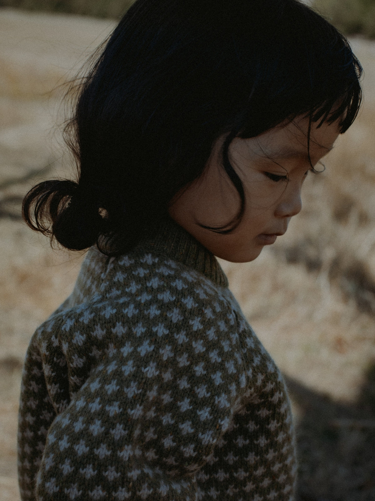 A close-up side profile of a young child with dark hair wearing a brown and white star-patterned knit sweater, set against a soft-focus outdoor background.