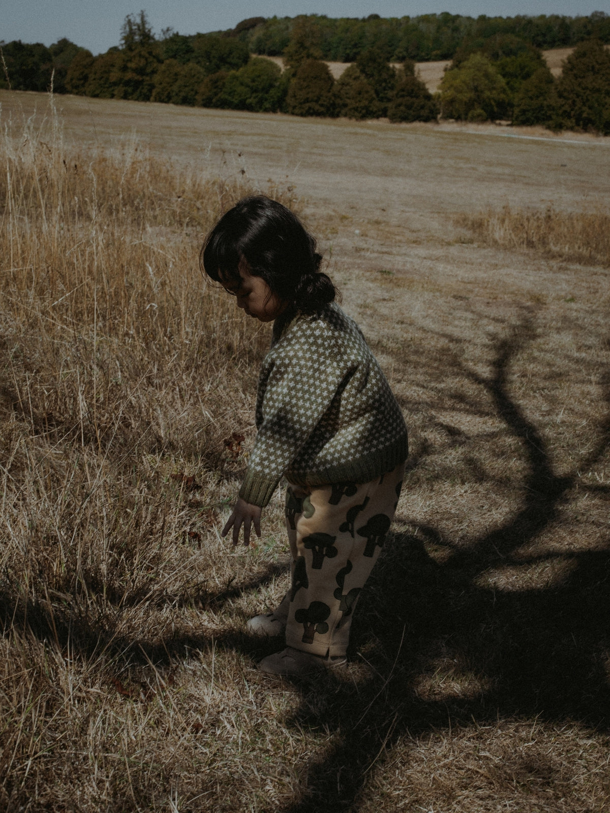 A child wearing a green and white patterned knit sweater and beige pants with black spots stands in a dry grassy field outdoors.