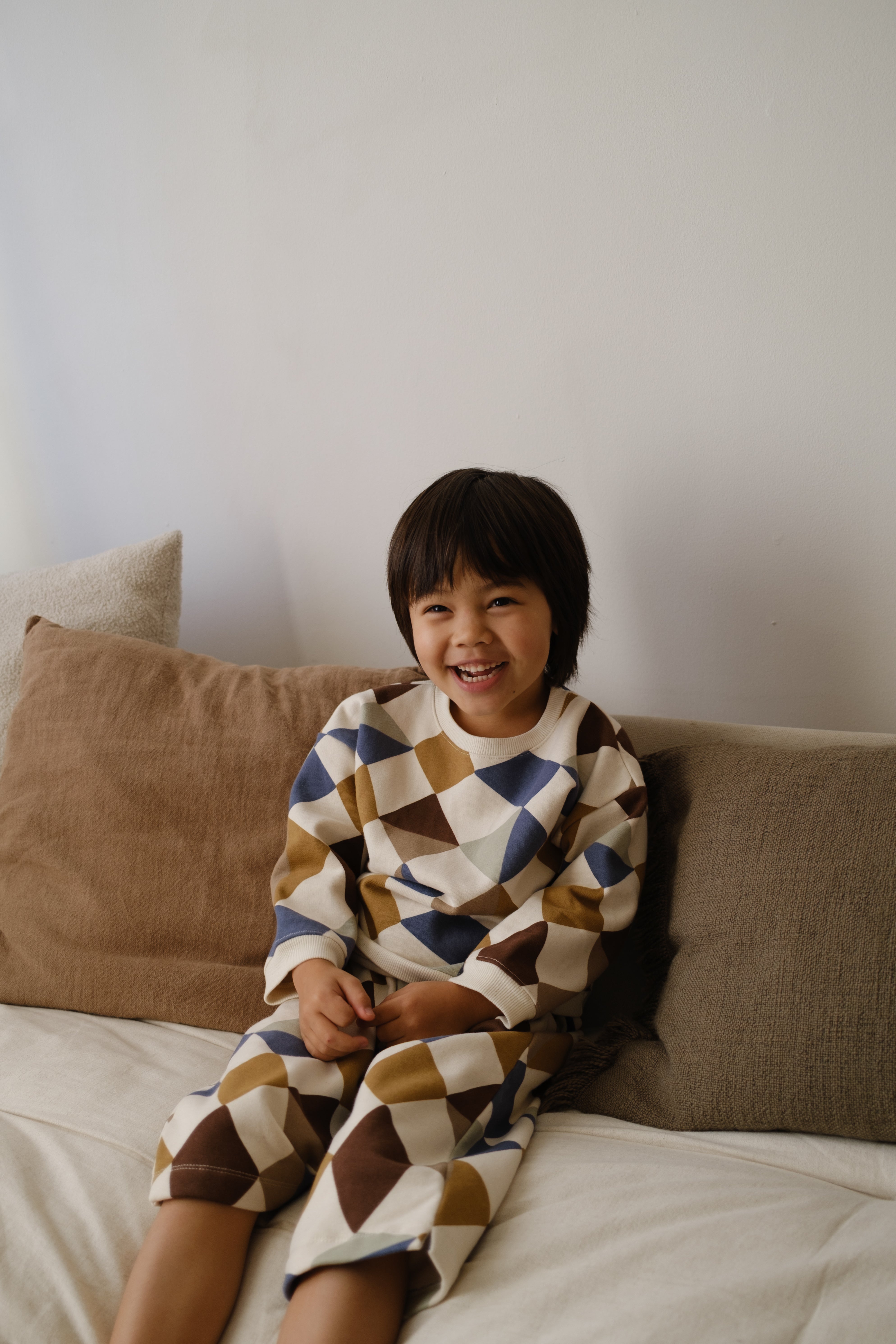 toddler in matching cream sweatshirt & pants with multicolour geometric patchwork, sitting on bed