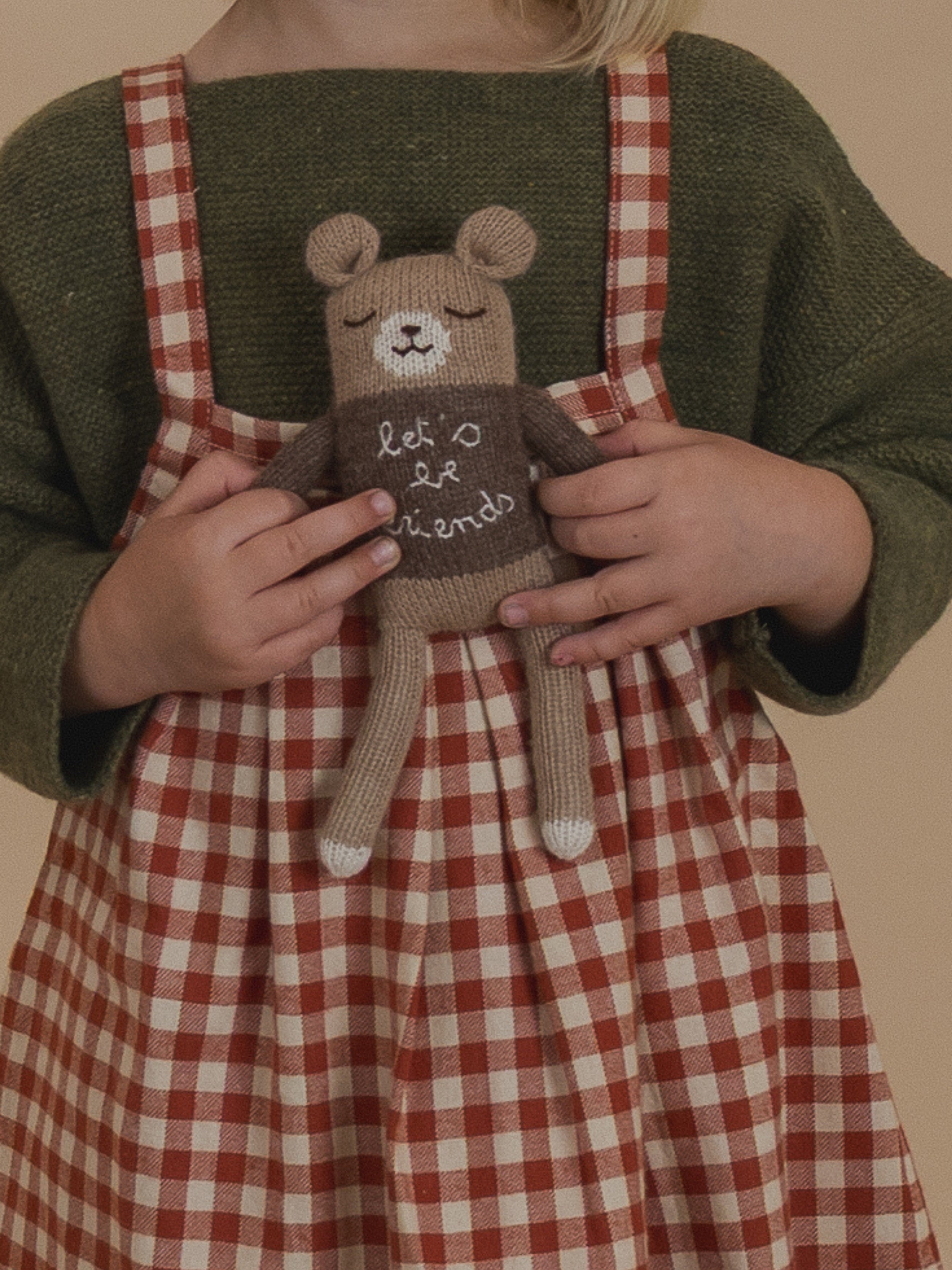 close up of child in wool jumper & gingham skirt, holding wool teddy toy with let's be friends jumper
