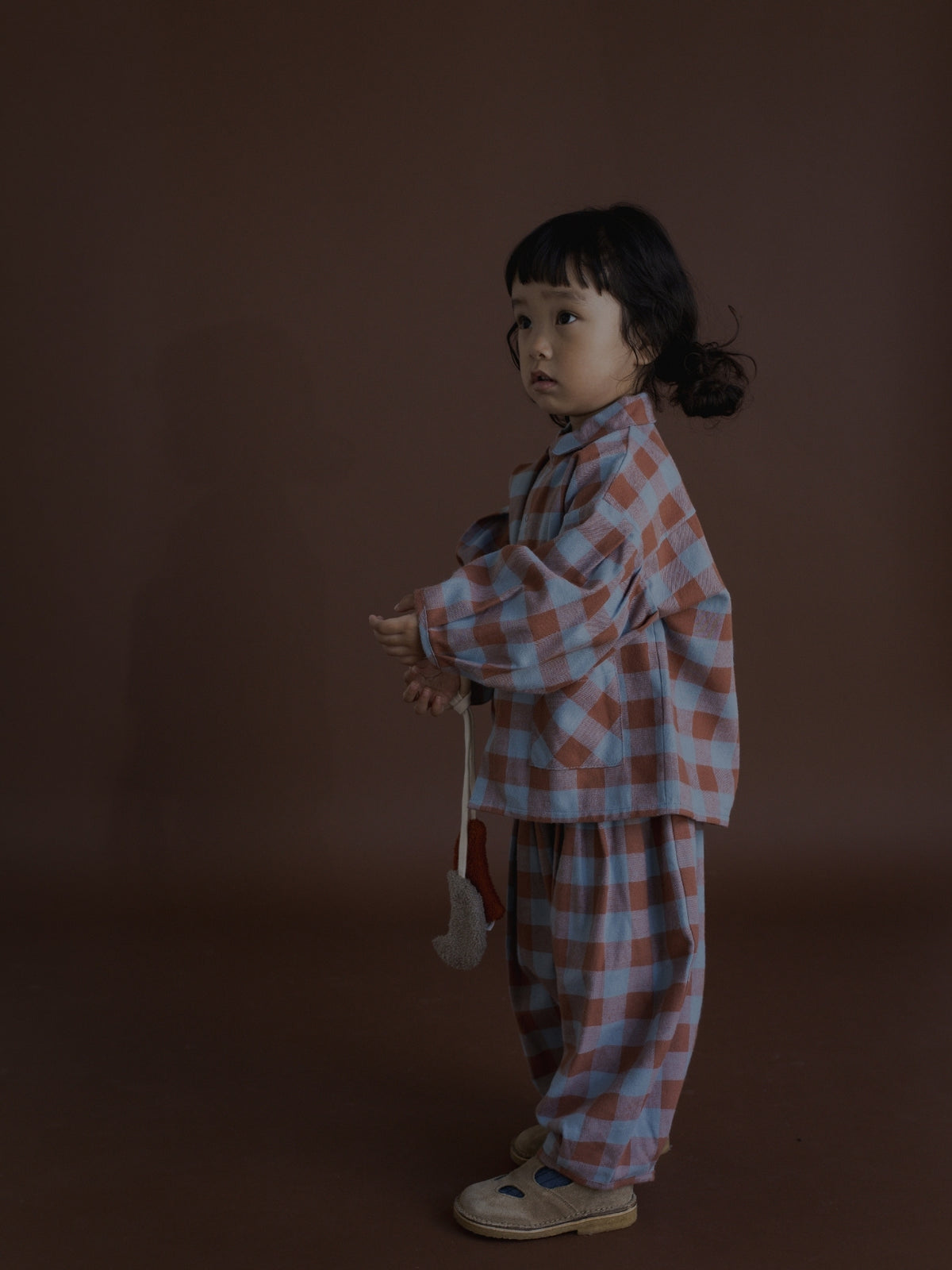 A young child stands in profile against a dark brown studio background, wearing a matching two-piece plaid outfit in muted pink, blue, and white, with light beige shoes.