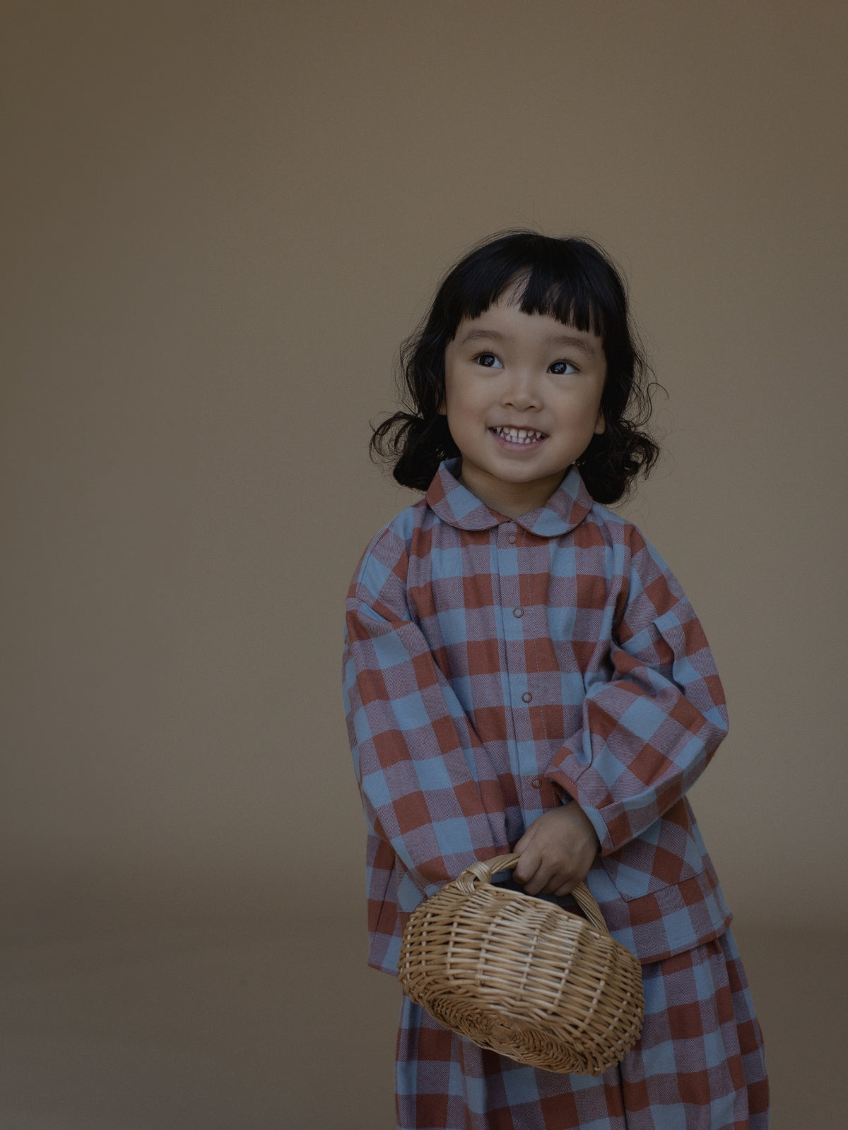 A young child with dark hair wearing a blue and rust-red plaid long-sleeve shirt, holding a small woven basket against a solid warm brown studio background.