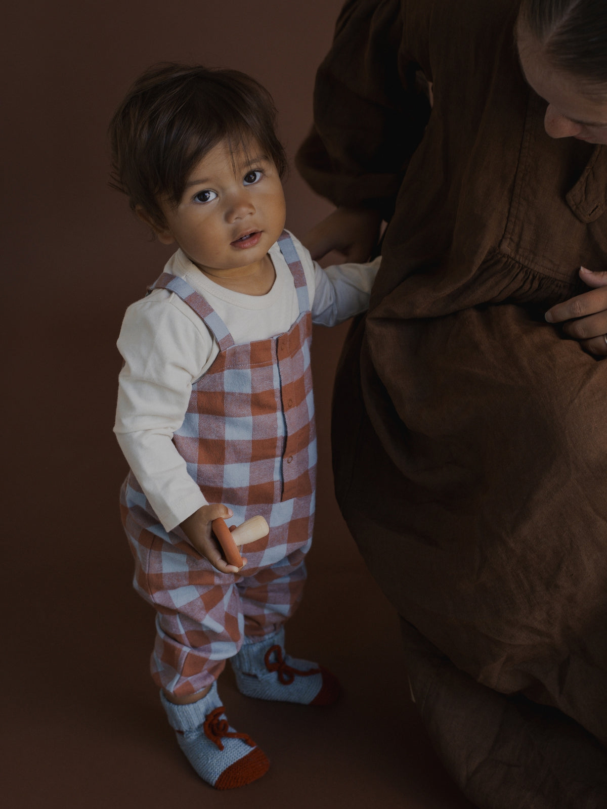 A toddler stands next to an adult in a studio setting, wearing a white long-sleeve shirt and brown-and-white gingham check overalls with light blue socks.