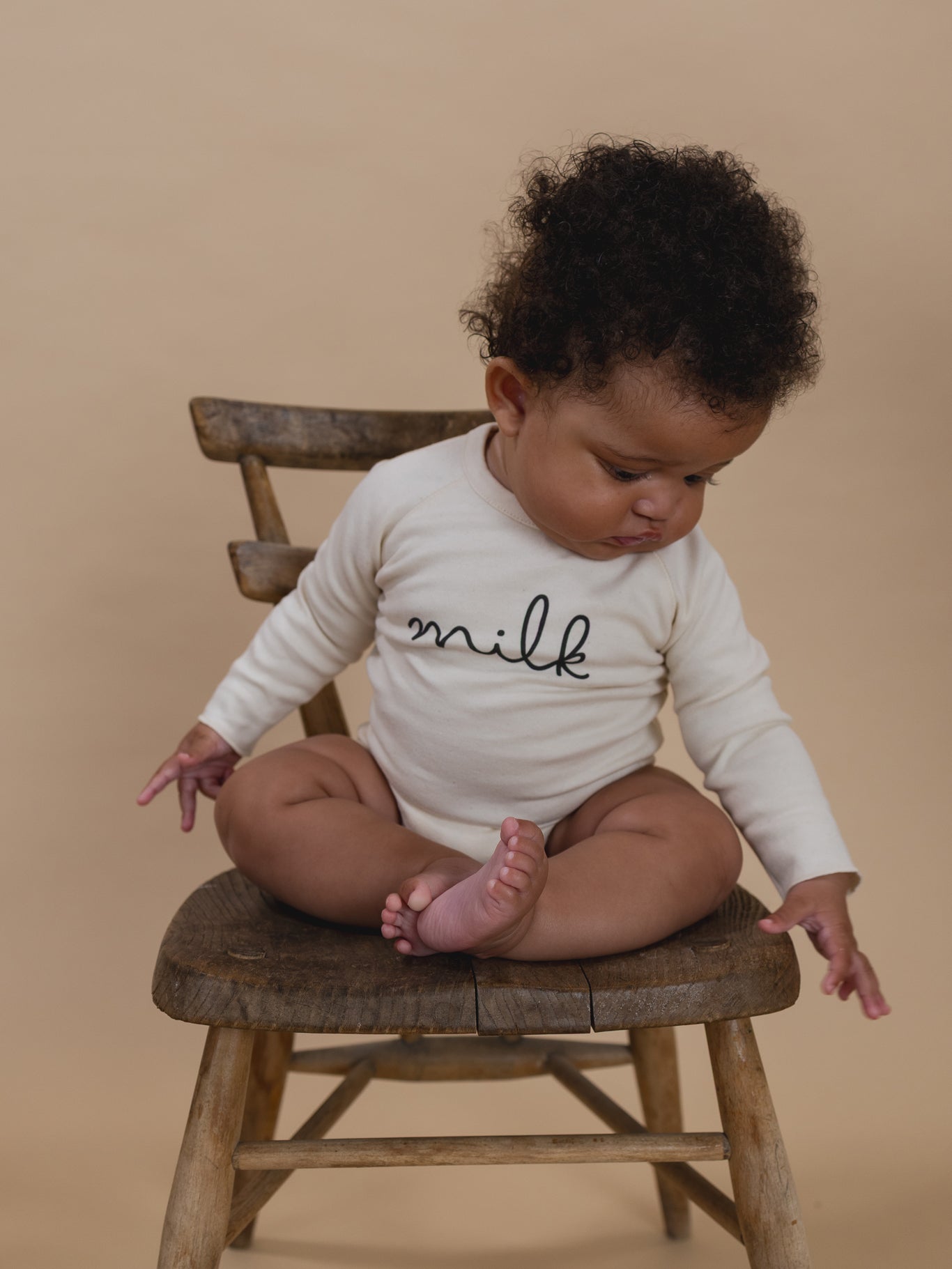 baby wearing cream undyed cotton bodysuit with milk text, sitting on a wooden chair
