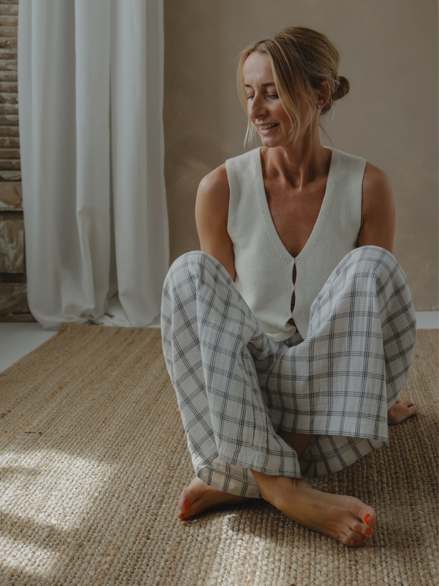 Woman sitting on a woven mat wearing a white sleeveless top and checkered pants in a softly lit room.