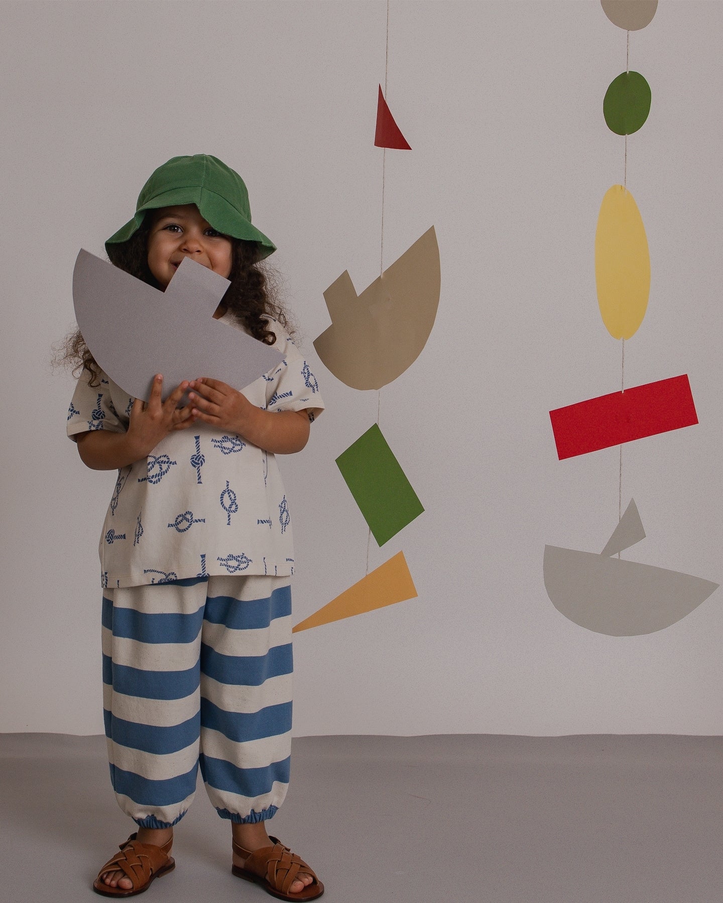 Child holding a paper boat in front of a colorful geometric wall hanging