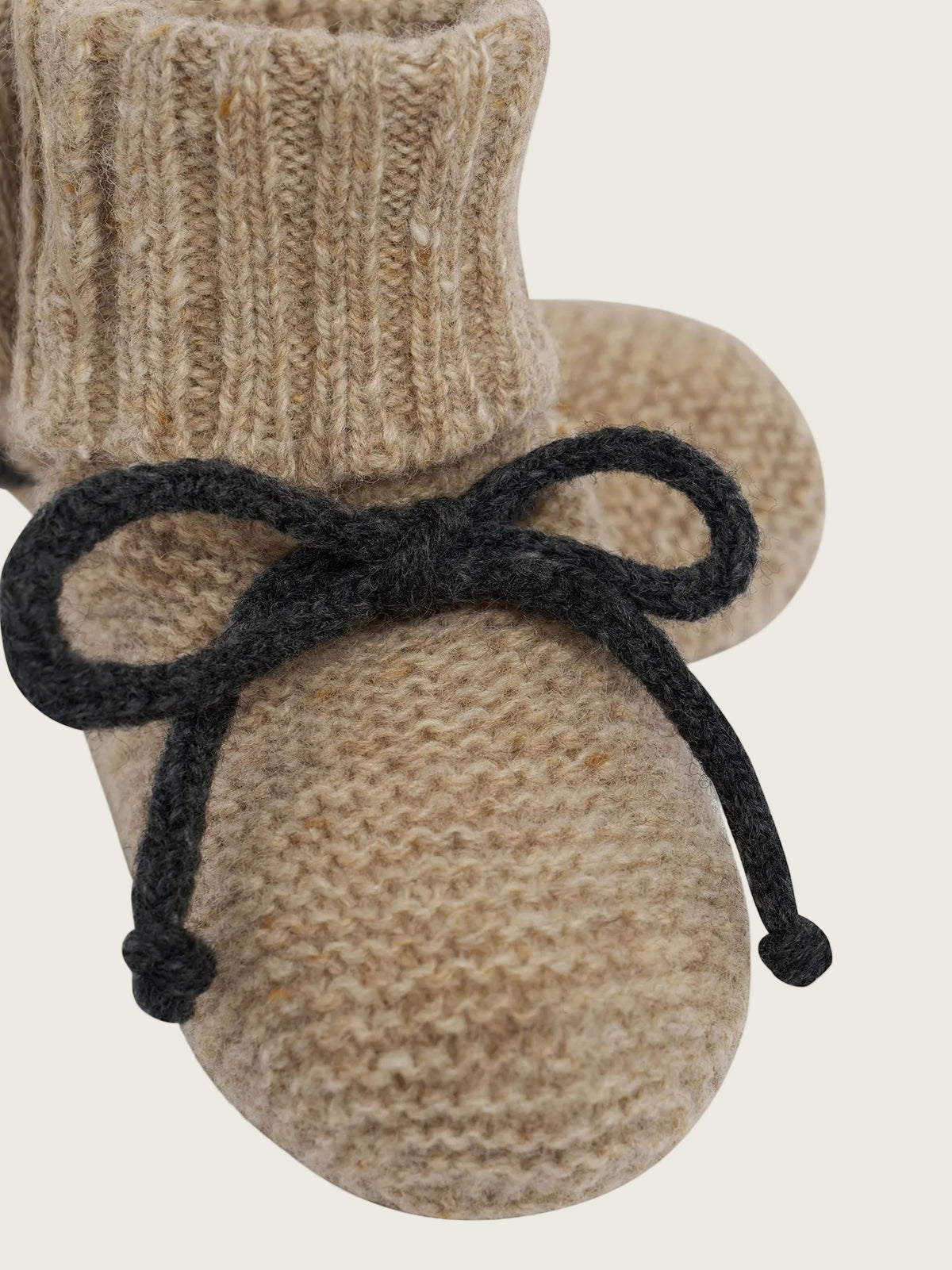 A pair of beige knit baby booties with a black yarn bow tied around the ankle, shown on a plain white background.