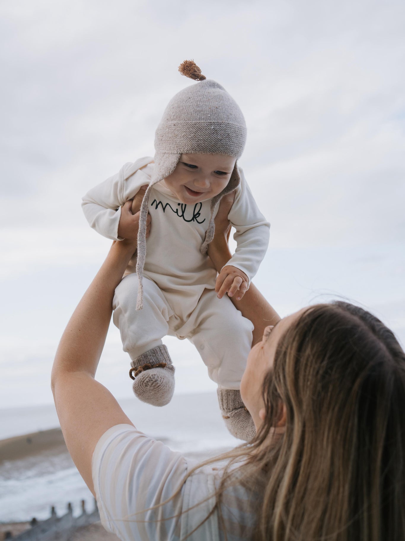 mum carrying baby in oatmeal wool booties with ribbons, with matching tassel hat and cream playsuit