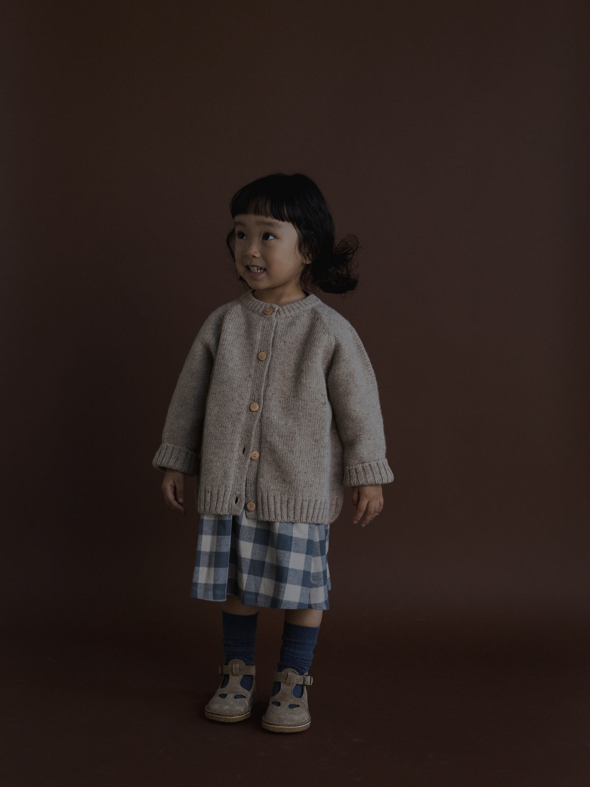 A toddler girl stands against a dark brown studio background wearing a light beige knit cardigan with wooden buttons, a blue and white plaid skirt, dark socks, and beige shoes.