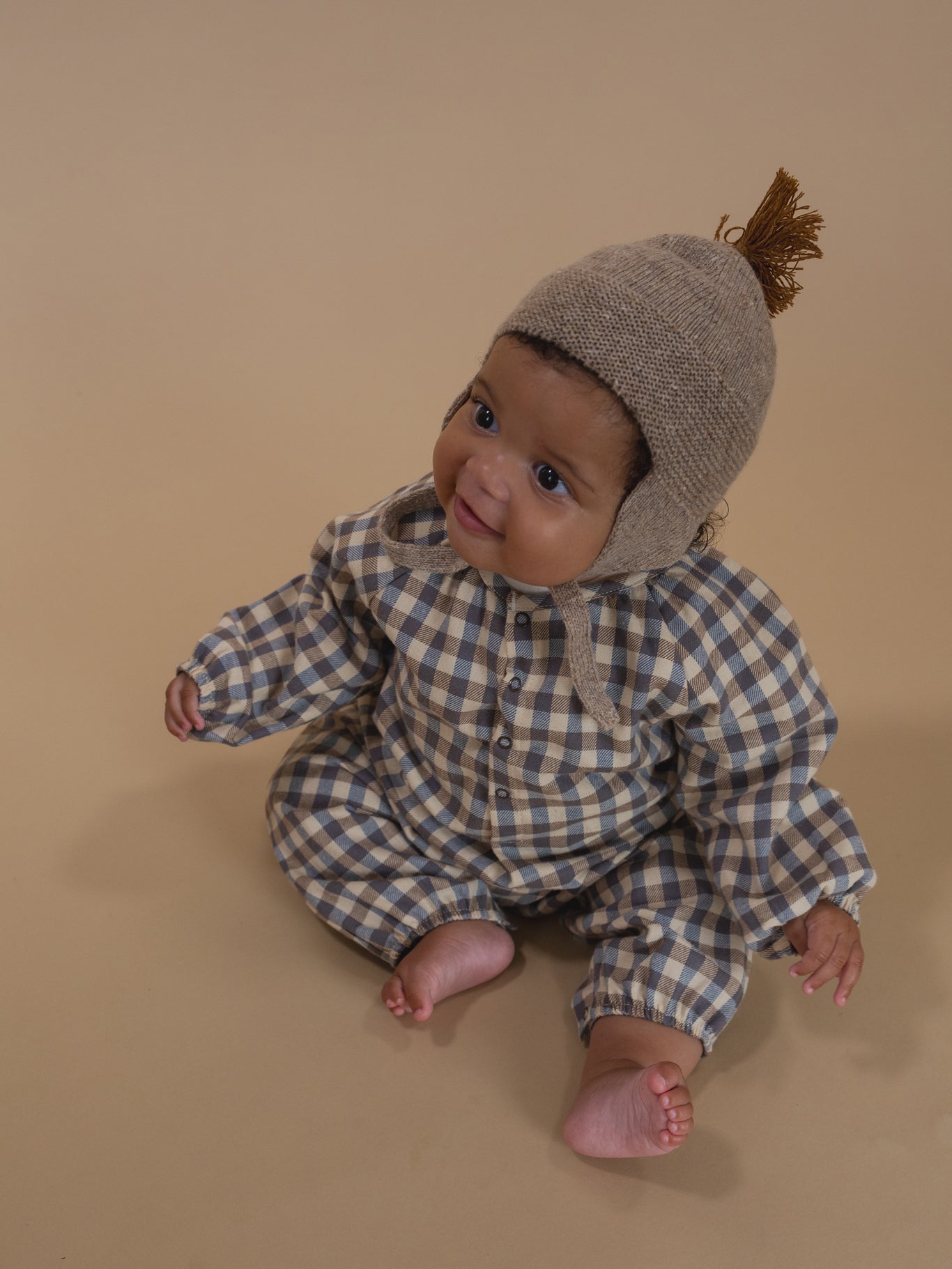 baby wearing oatmeal wool hat with brown tassel and chin tie, and gingham onesie, sitting on floor