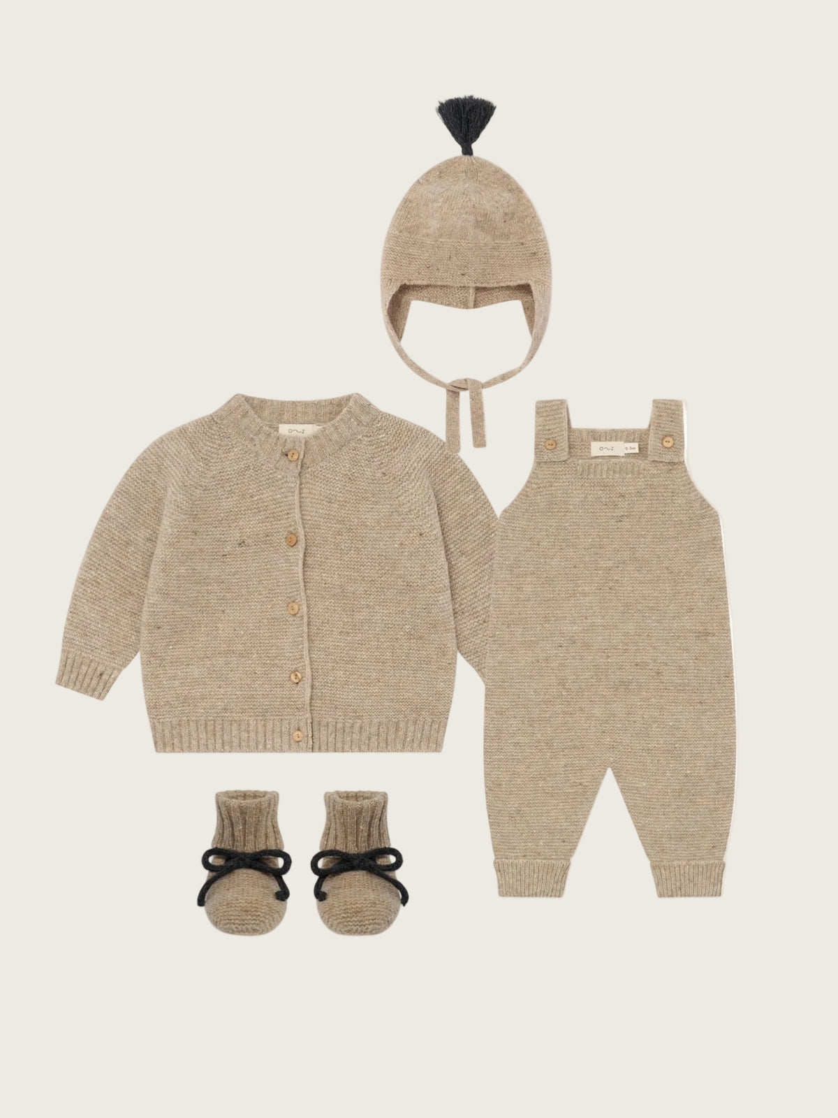 A baby knit clothing set in a warm beige/tan color, including a cardigan, overalls, a bonnet with a black pom-pom, and booties with black bows, arranged on a plain white background.
