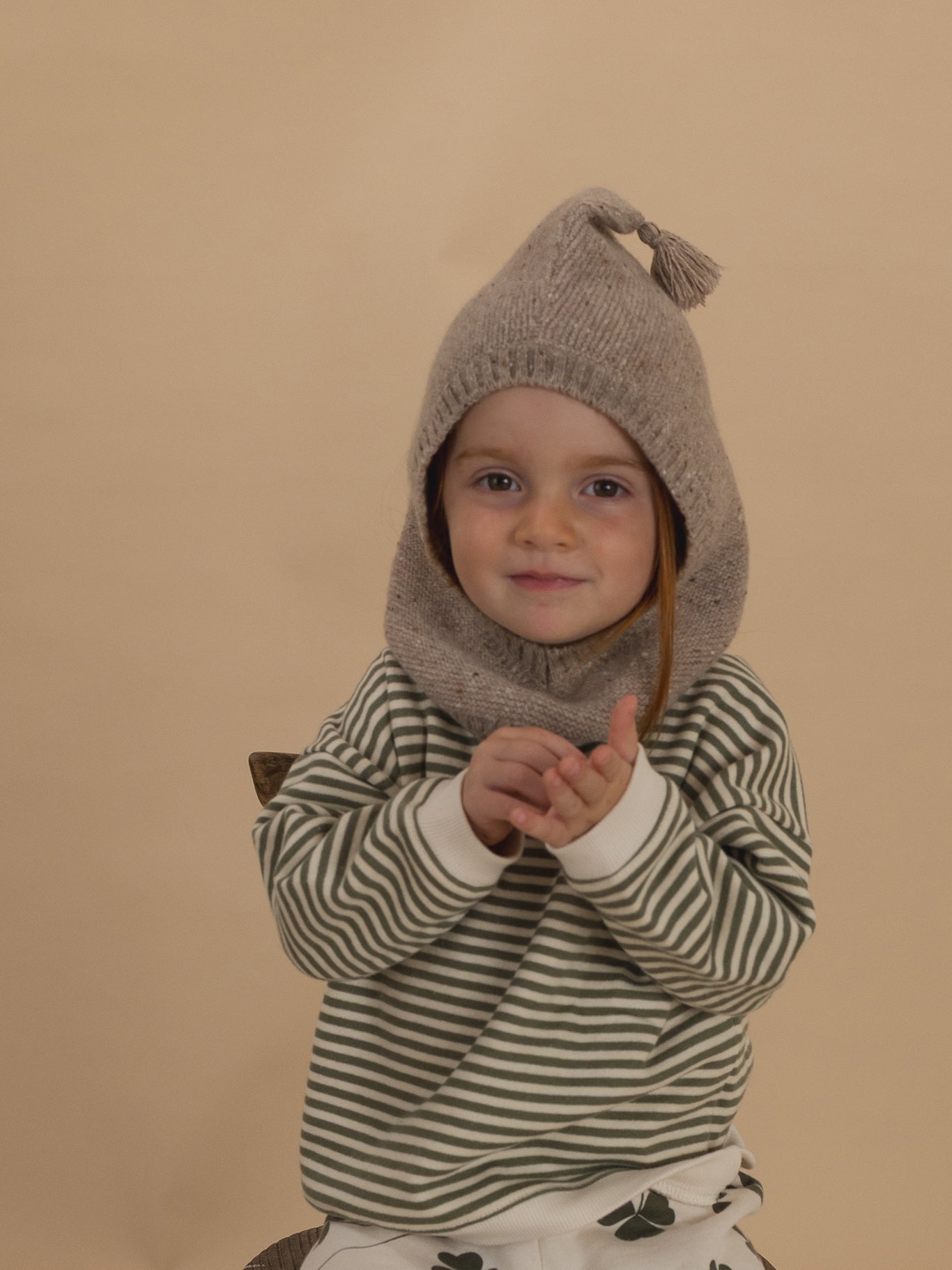close up of a toddler in oatmeal wool knitted balaclava, green striped sweatshirt and clover pants